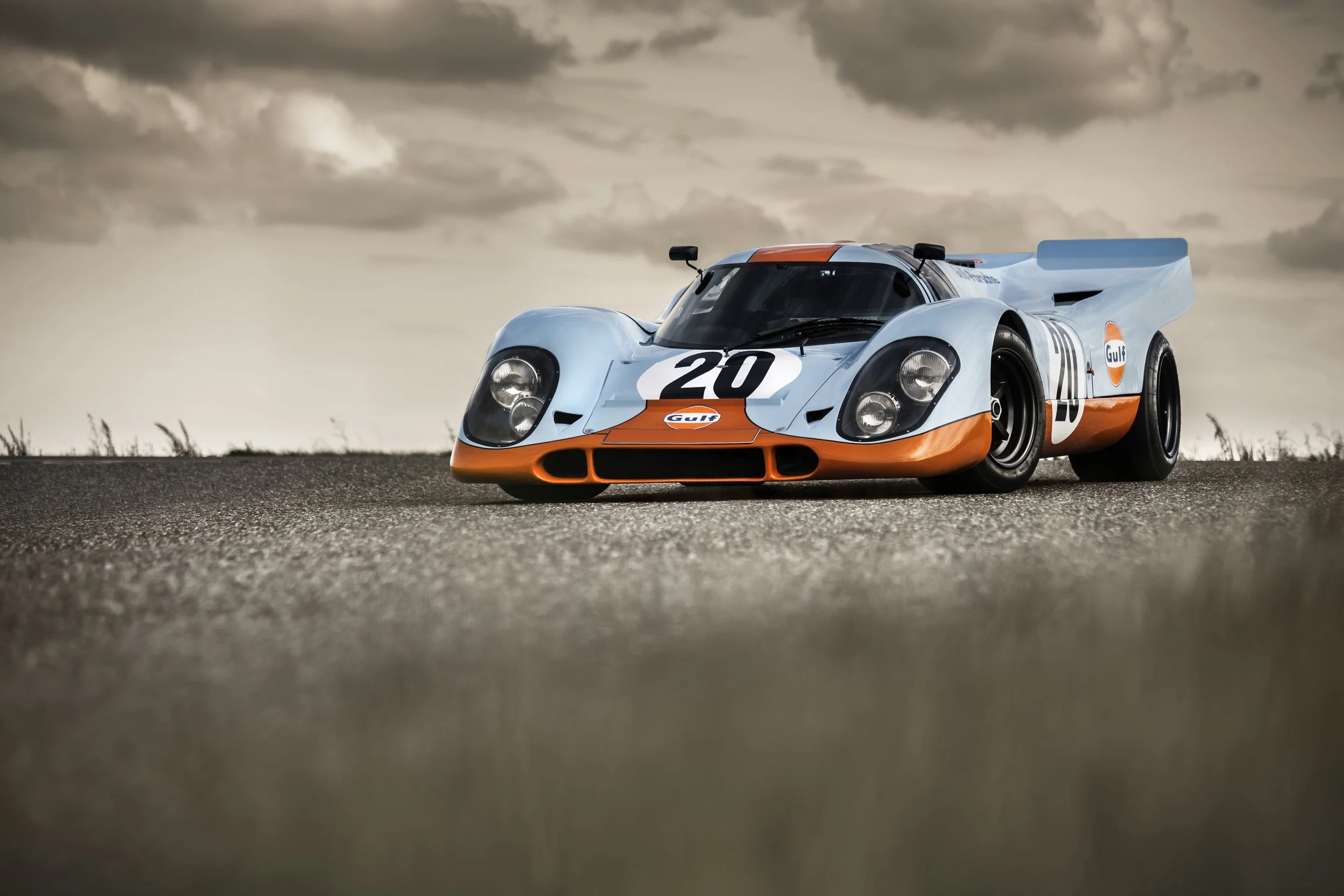 Ein orange-blauer Rennwagen mit der Nummer 20 und Gulf-Logo auf einer Straße bei bewölktem Himmel.