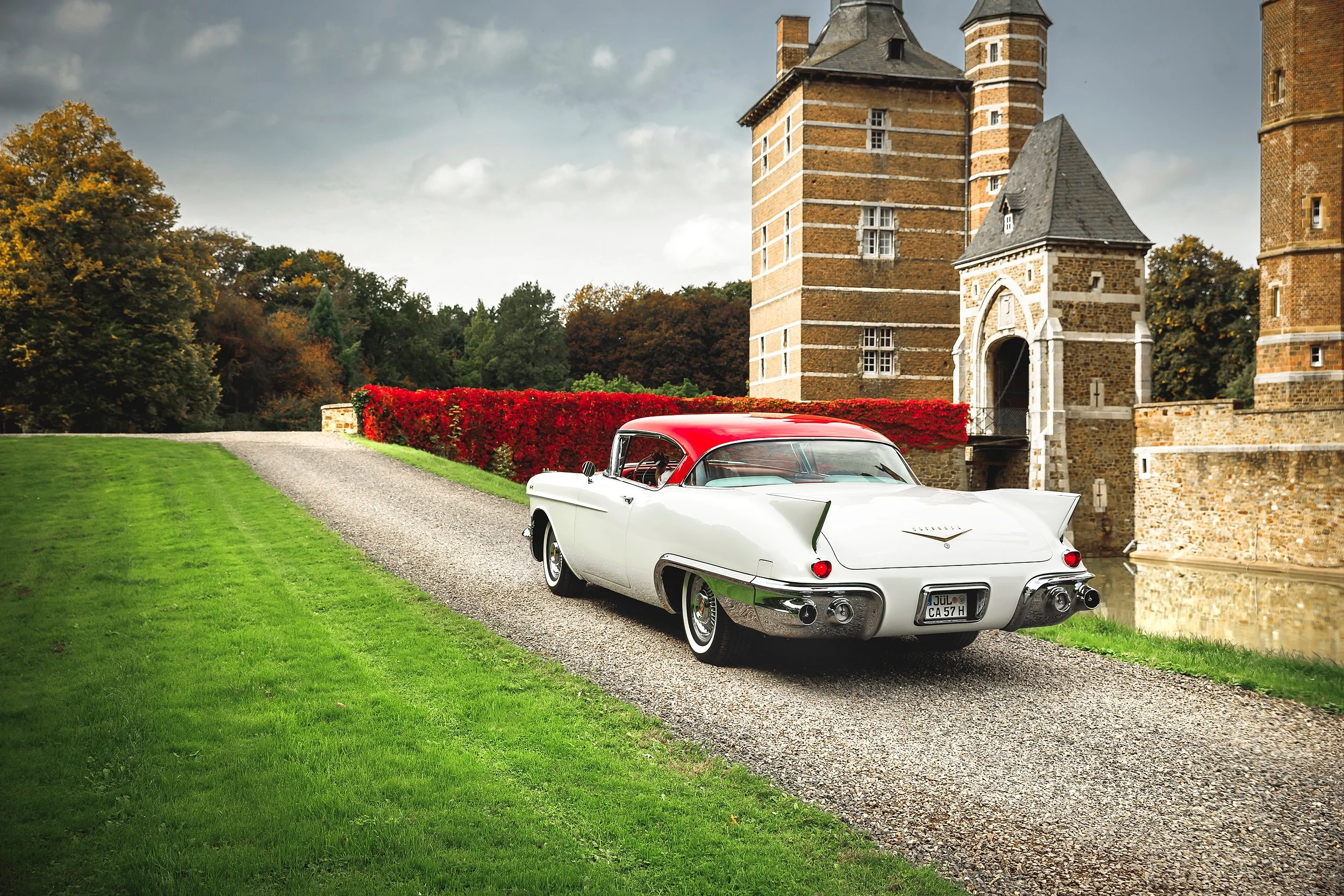 Ein weißer Oldtimer mit rotem Dach parkt an einem Teich vor einem historischen Schloss mit Türmen, umgeben von Bäumen und einem gepflegten Garten.