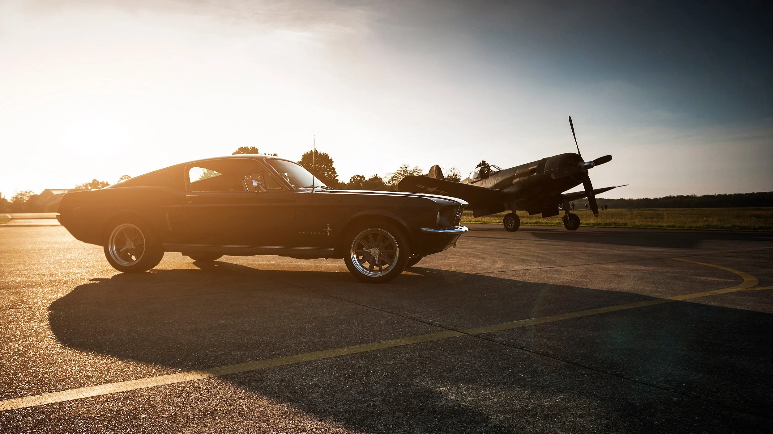 Ein schwarzer Ford Mustang parked auf einem Flugplatz bei Sonnenuntergang, im Hintergrund ein abgestelltes Flugzeug.