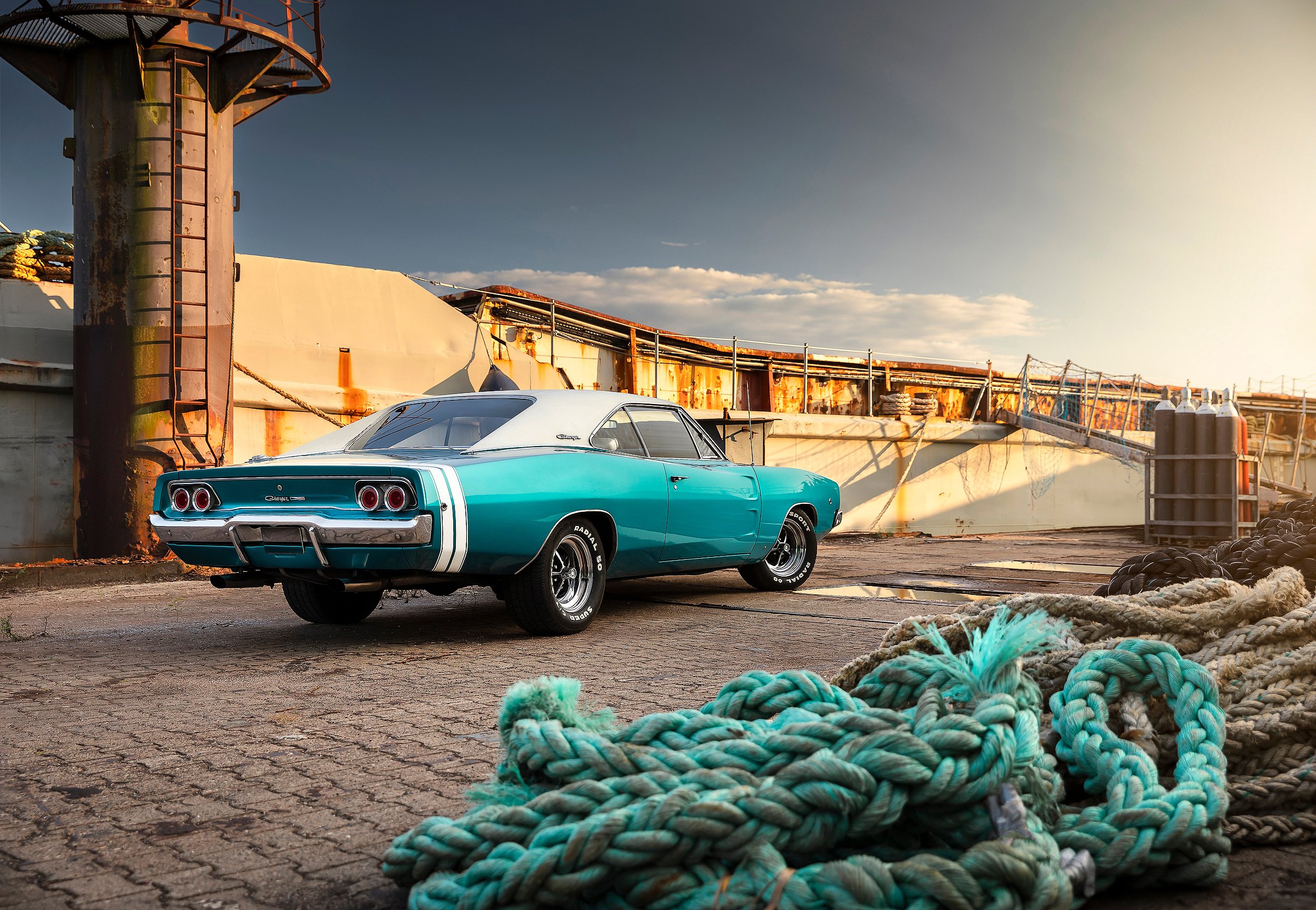 Ein blauer Muscle-Cars-Auto steht vor einem alten, rostigen Frachtschiff am Kai, mit Kletterseilen an der Seite. Im Vordergrund liegen verdrehte, farbige Seile auf dem Boden.