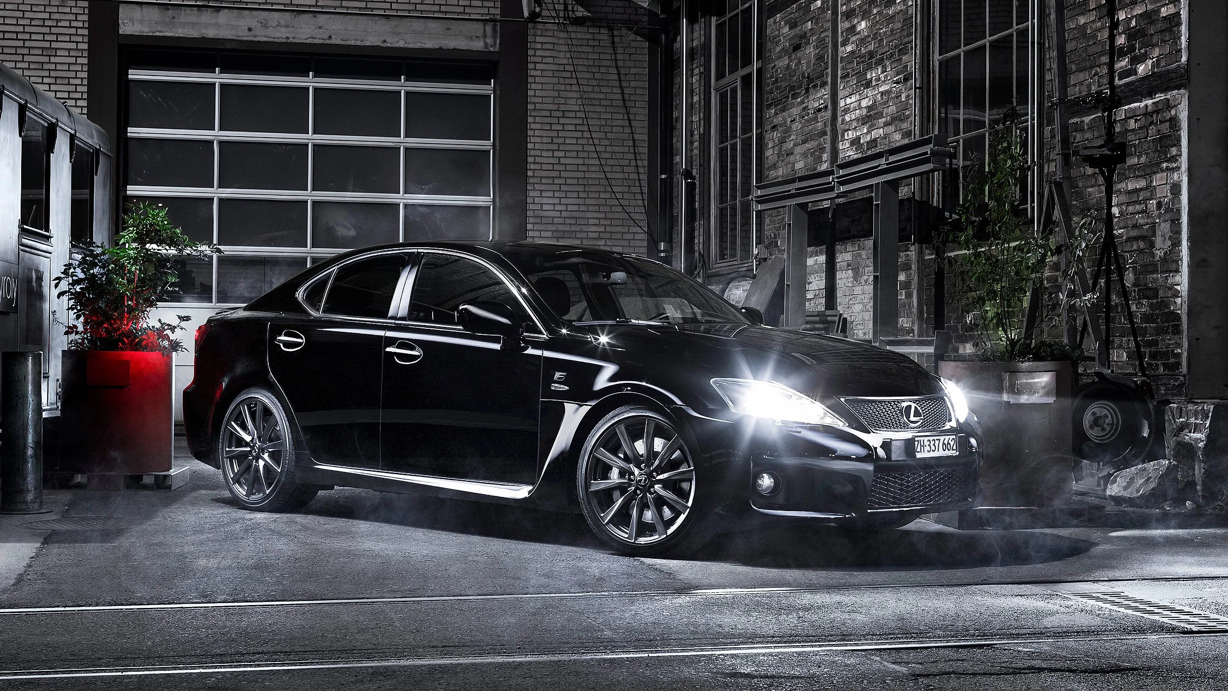 Ein schwarzer Lexus-Auto parkt in einer urbanen, nächtlichen Umgebung neben einem Gebäude mit Backsteinmauern und großen Fenster.