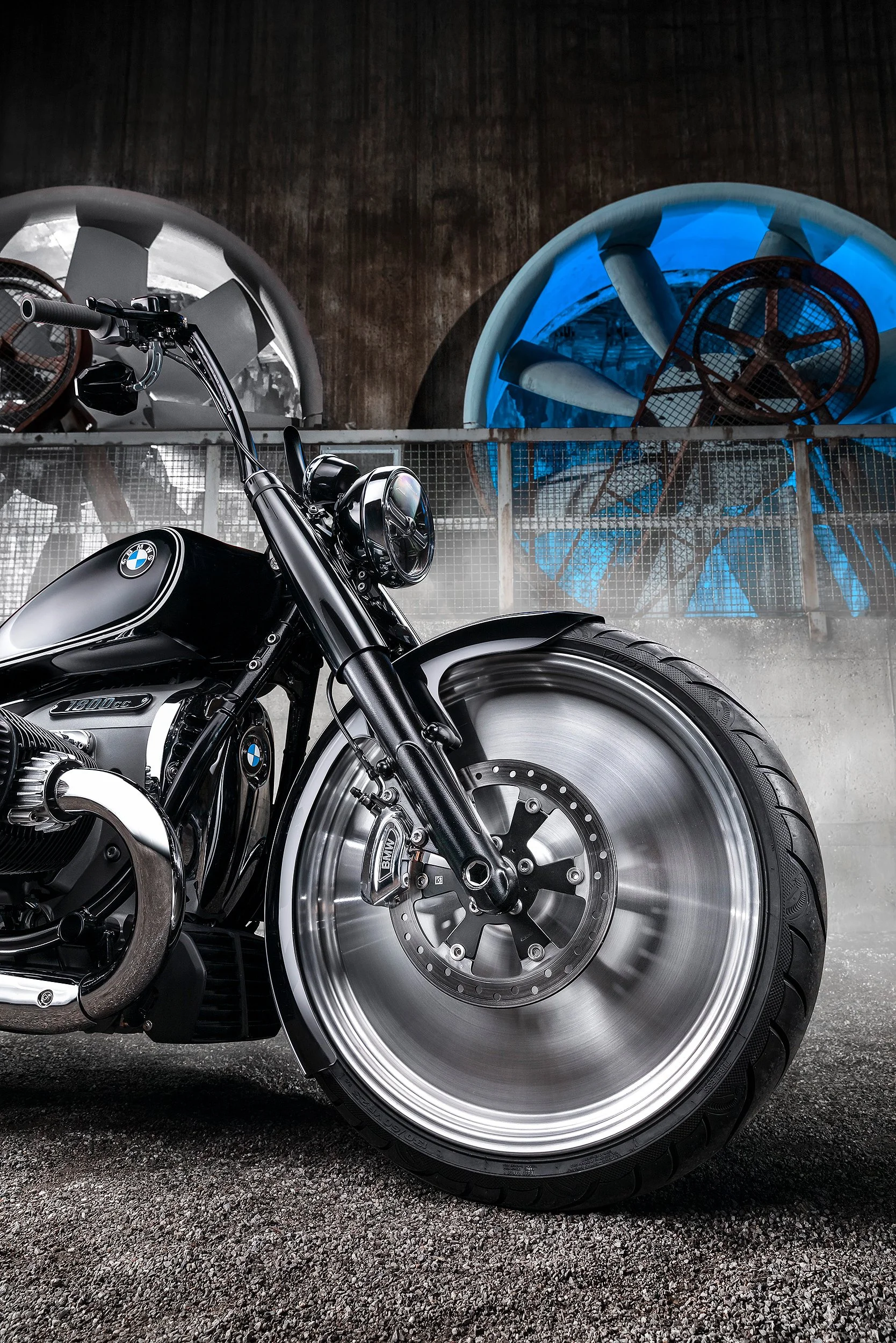 Nahaufnahme eines schwarzen BMW Motorrads mit hellem, glänzendem Felgenrad und großen Reifen, vor einer Wasseraufbereitungsanlage mit großen blauen und grauen Zisternen.