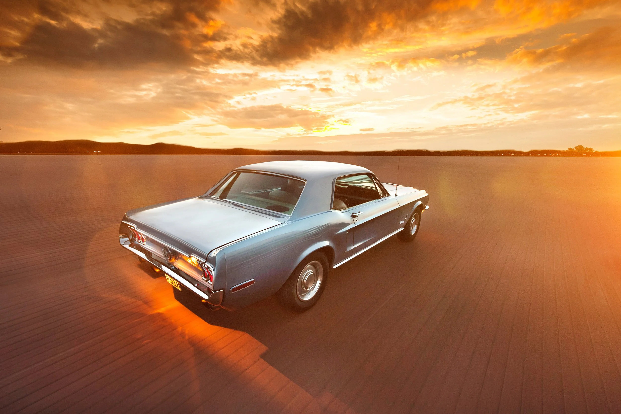 Ein klassischer silberner Oldtimer-Auto fährt auf einer Brücke bei Sonnenuntergang, mit einem dramatischen Himmel voller Wolken und orangefarbenem Licht im Hintergrund.
