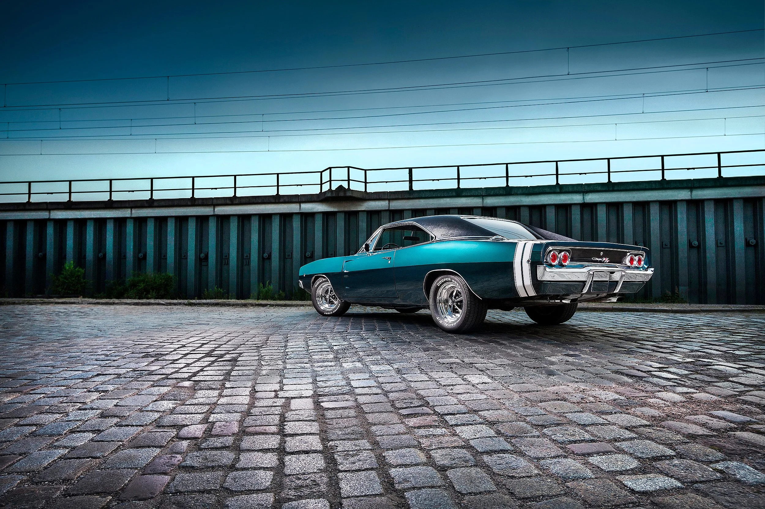 Ein klassischer schwarzer Muscle-Car, ein Chevrolet Chevelle, parkt auf einer gepflasterten Straße vor einer hohen grünen Metallverkleidung, im Hintergrund ein blauer Himmel mit wenigen Wolken.
