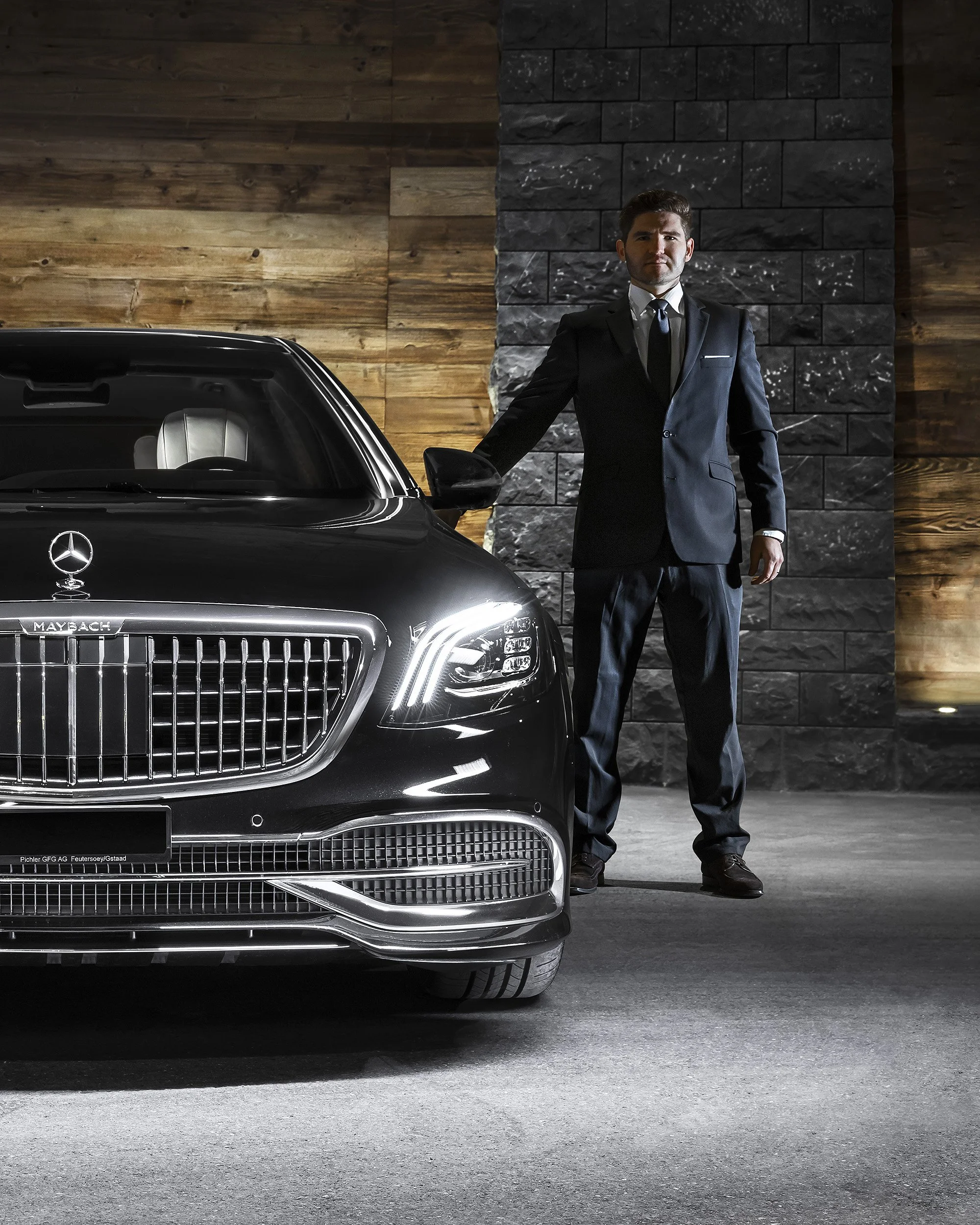 Ein Mann in einem schwarzen Anzug mit Krawatte steht neben einem schwarzen Mercedes-Benz Fahrzeug in einem Raum mit Holzwand und Steinwand. Er hält die Autotür am Griff.