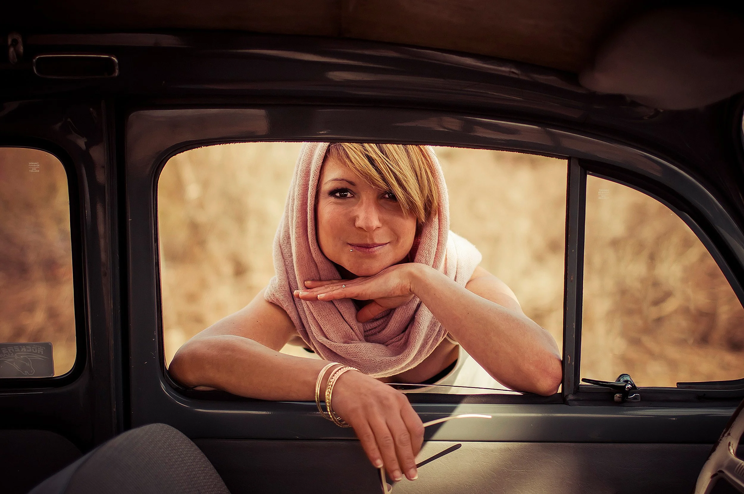 Junge Frau mit kurzen roten Haaren, pinkem Schal und Piercing, blickt freundlich aus einem Autofenster, umgeben von einer natürlichen, herbstlichen Umgebung.