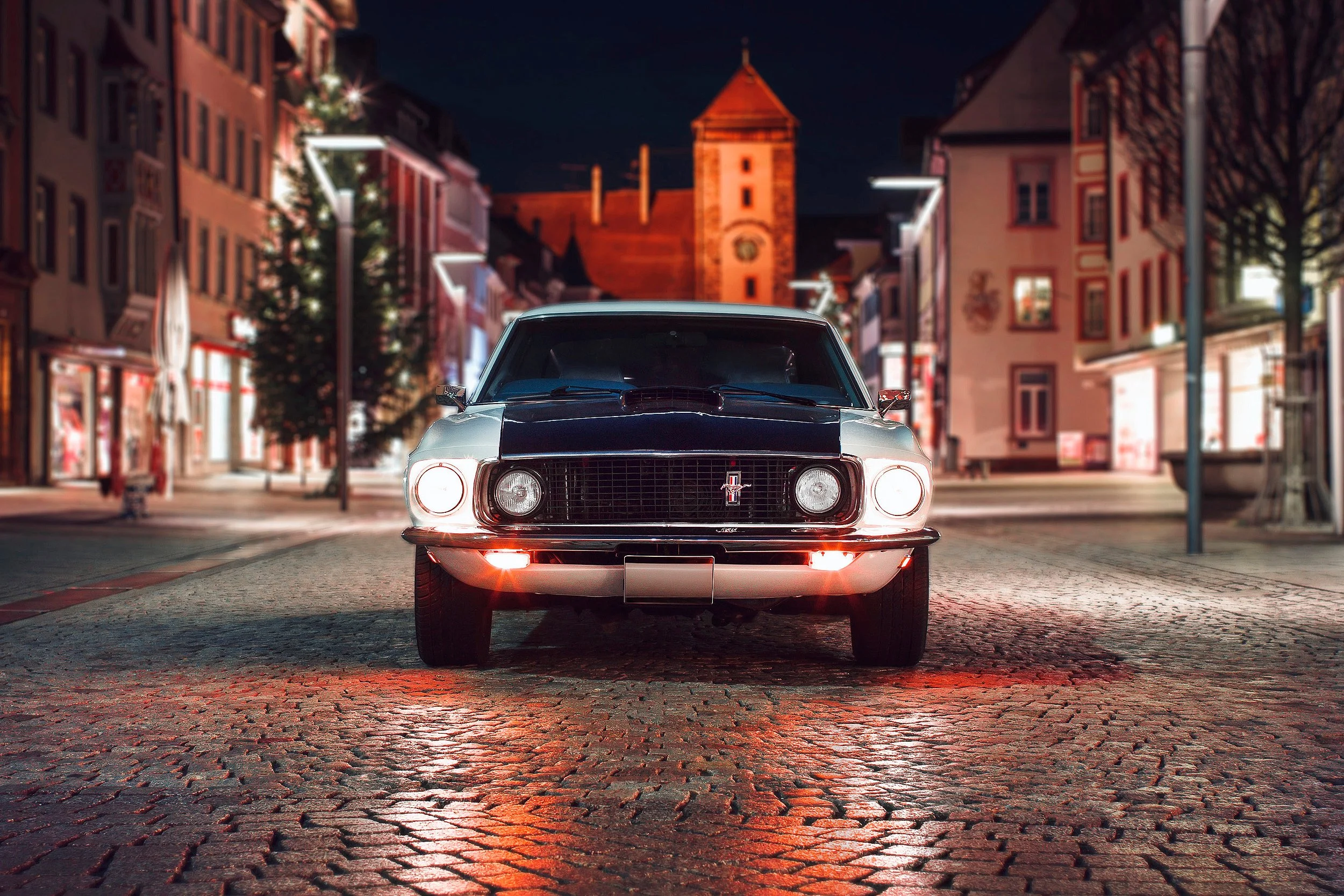 Ein schwarzer Oldtimer, ein Ford Mustang, steht auf einer gepflasterten Straße in einer Stadt bei Nacht. Im Hintergrund sind mehrstöckige Gebäude mit beleuchteten Fenstern und ein Kirchturm sichtbar. Die Straße ist beleuchtet und es ist dunkel.