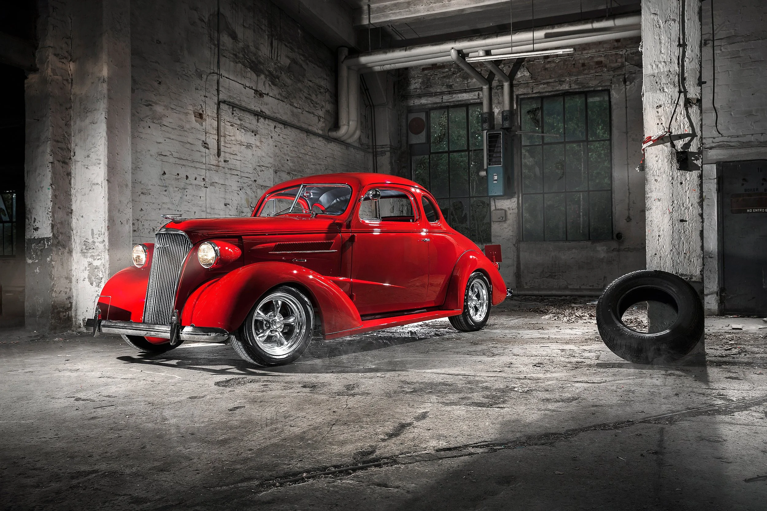 Ein rotes Vintage-Auto in einer alten, verfallenen Lagerhalle mit großen Fenstern und sichtbaren Rohren an der Decke.