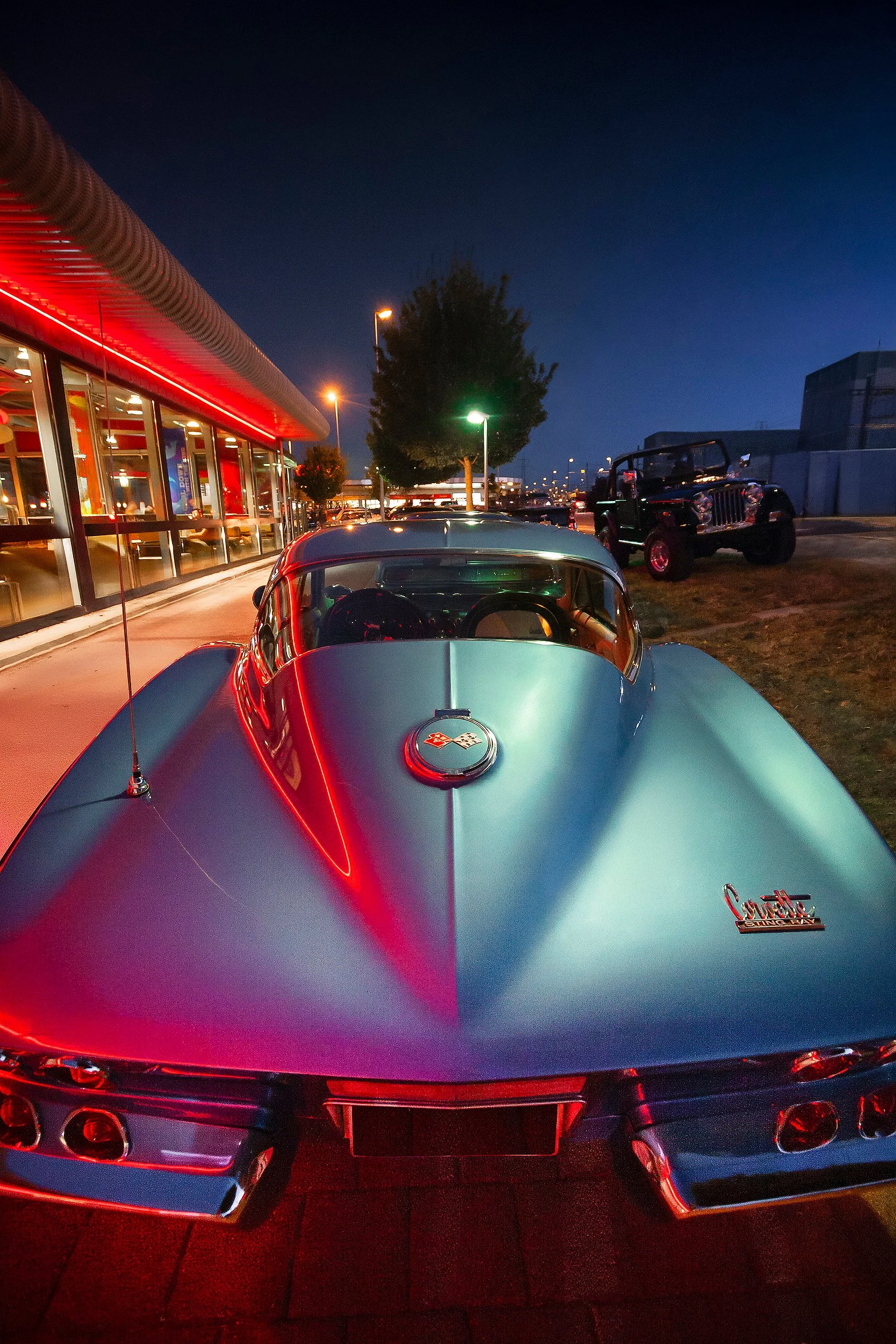 Ein silberner Chevrolet-Corvette mit rotem Neonlicht an der Decke und einem dunklen Jeep bei Nacht auf einem Parkplatz.