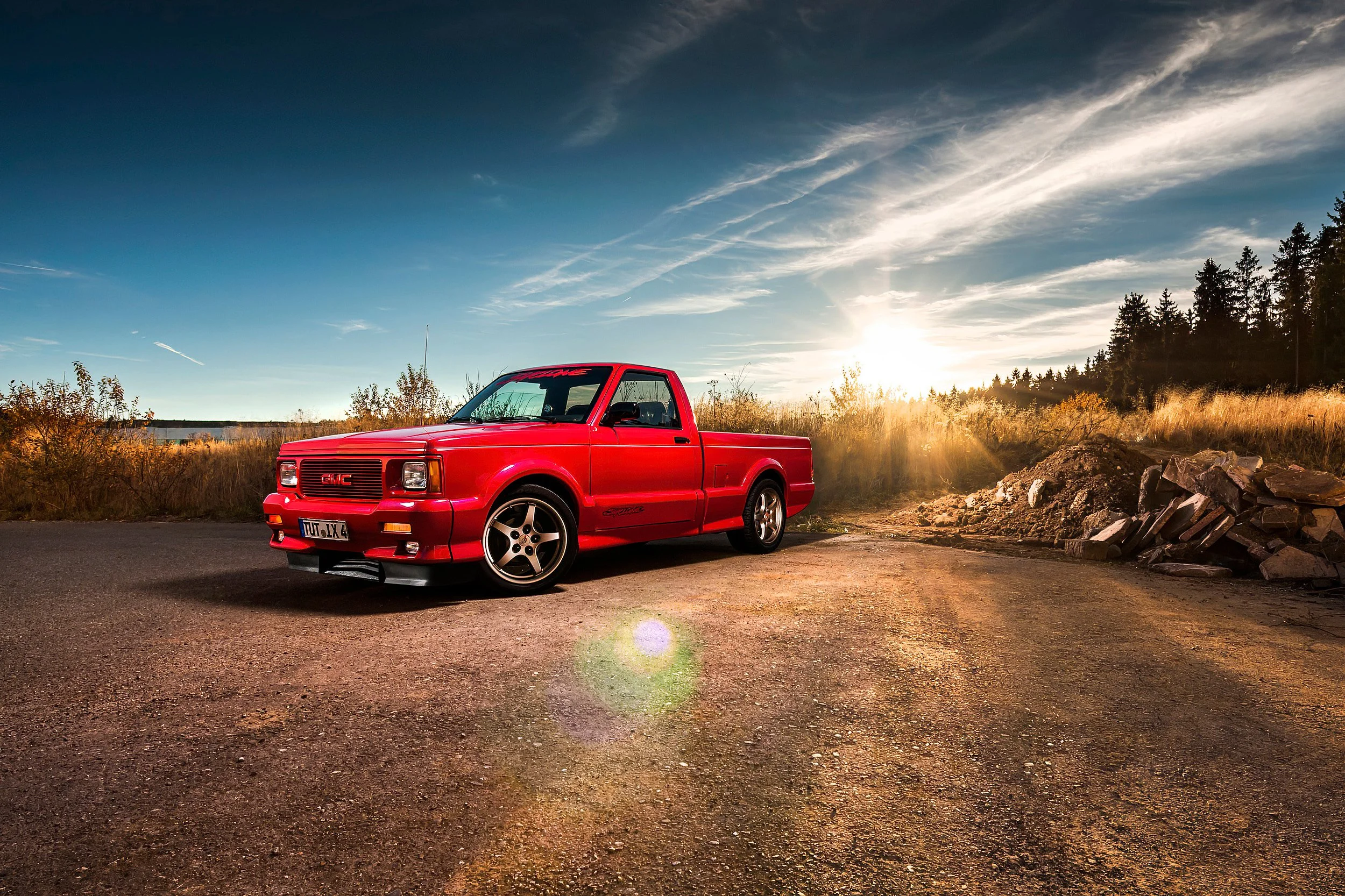Ein roter GMC-Canyon Pickup-Truck steht auf einer unbefestigten Straße bei Sonnenuntergang, im Hintergrund Bäume und ein Himmel mit leichten Wolken.