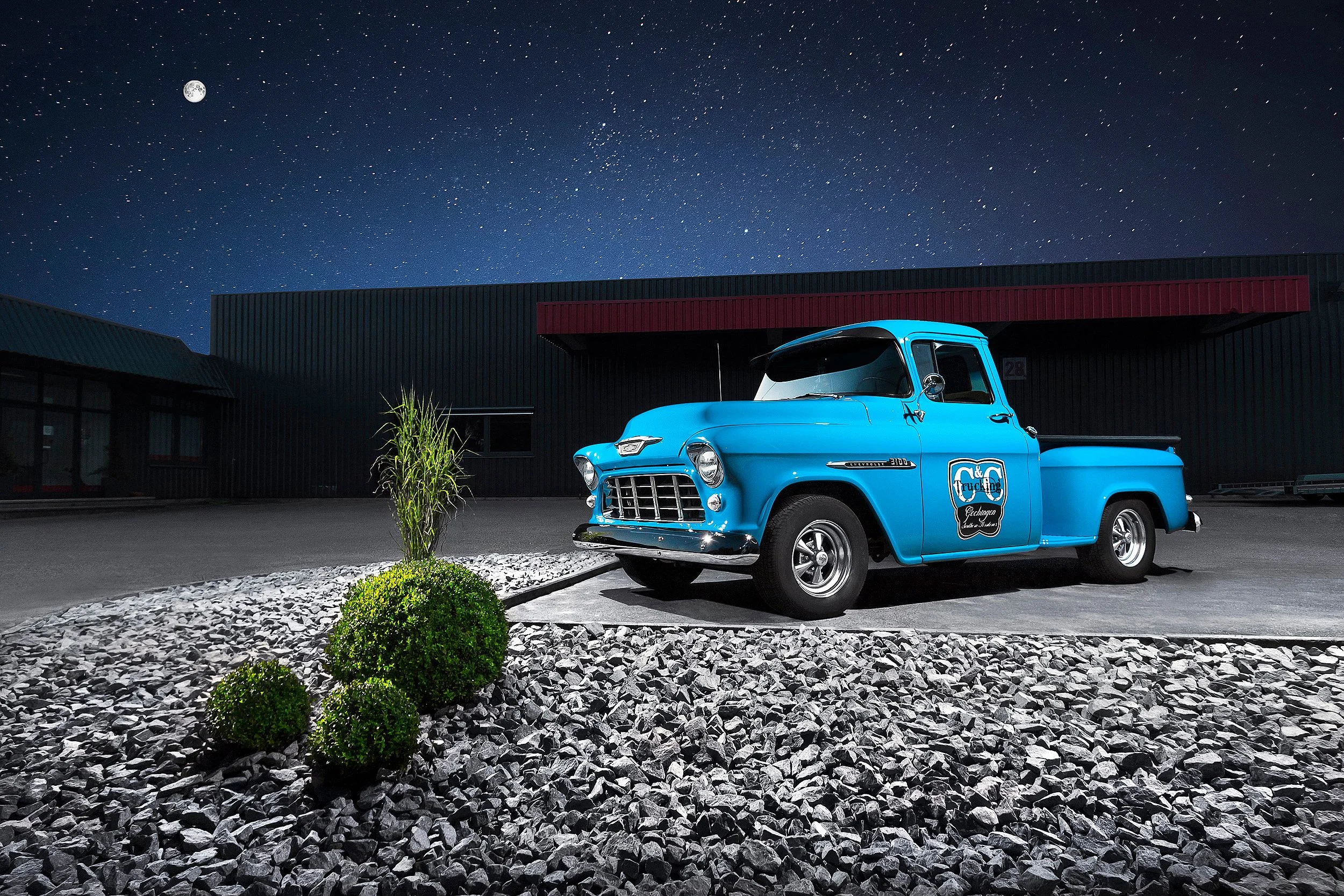 Ein alter blauer Pick-up Truck steht in einer dunklen Umgebung bei Nacht, unter einem sternenklaren Himmel mit Vollmond. Es gibt einige Sträucher und eine kleine Pflanze im Vordergrund.