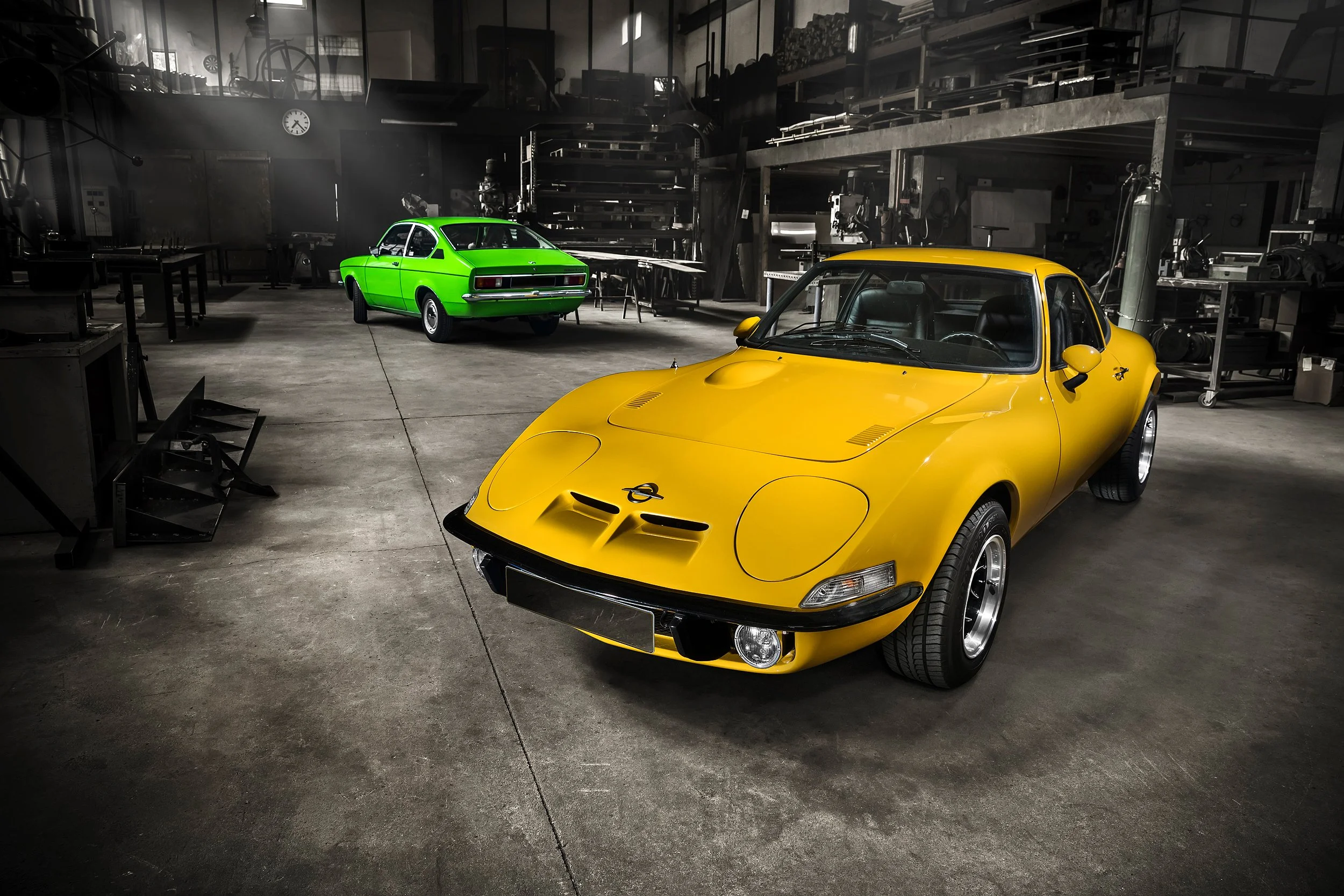 Zwei bunte Oldtimer-Autos, ein gelber Sportwagen im Vordergrund und ein grüner Pkw im Hintergrund, stehen in einer Werkstatt oder Garage mit Werkzeugen und Regalen an den Wänden.