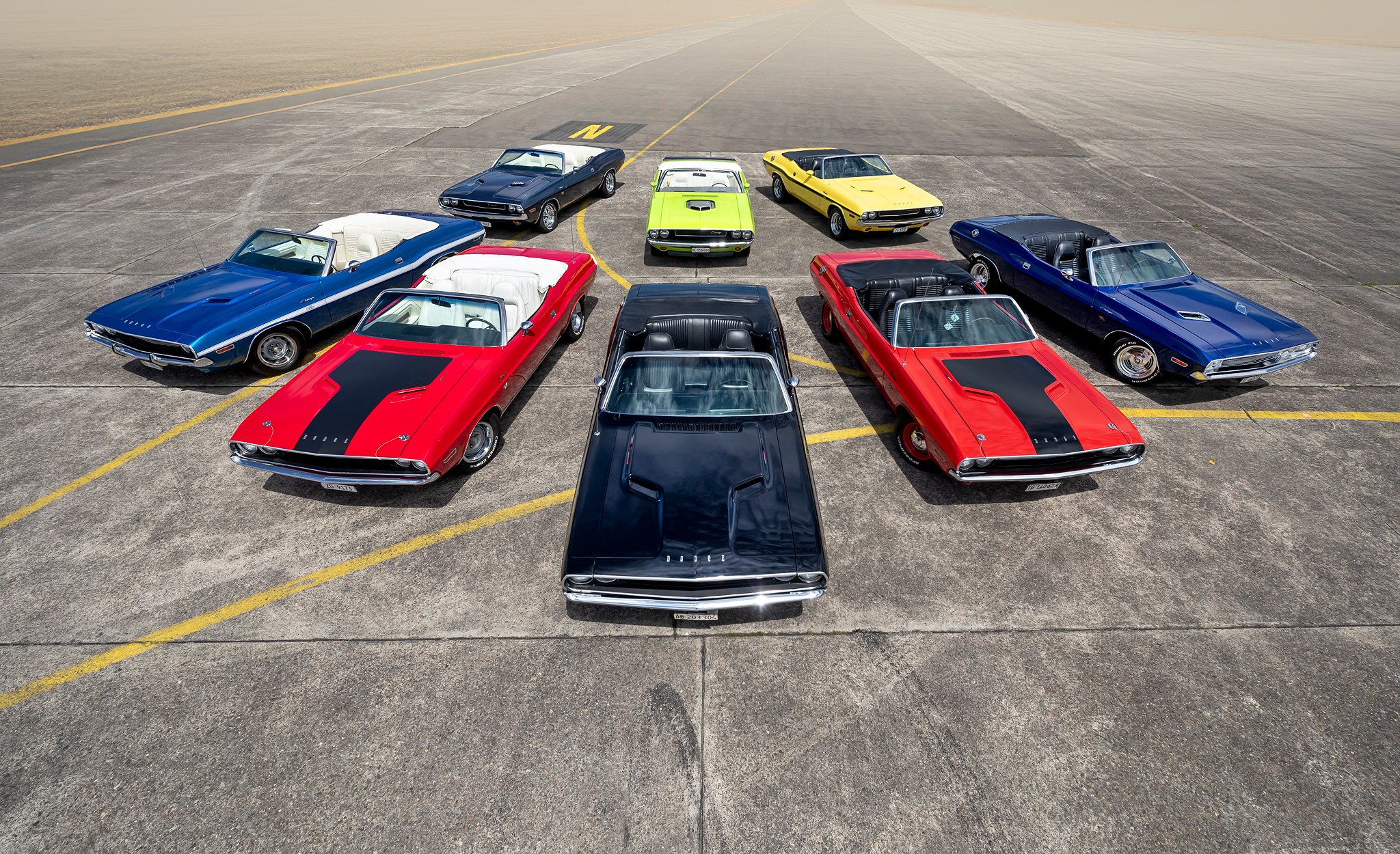 Zehn klassische amerikanische Muscle Cars auf einem Flugfeld, darunter Dodge, Chevrolet und Ford, mit flacher Perspektive und in einer Anordnung, die an eine Auto-Show erinnert.
