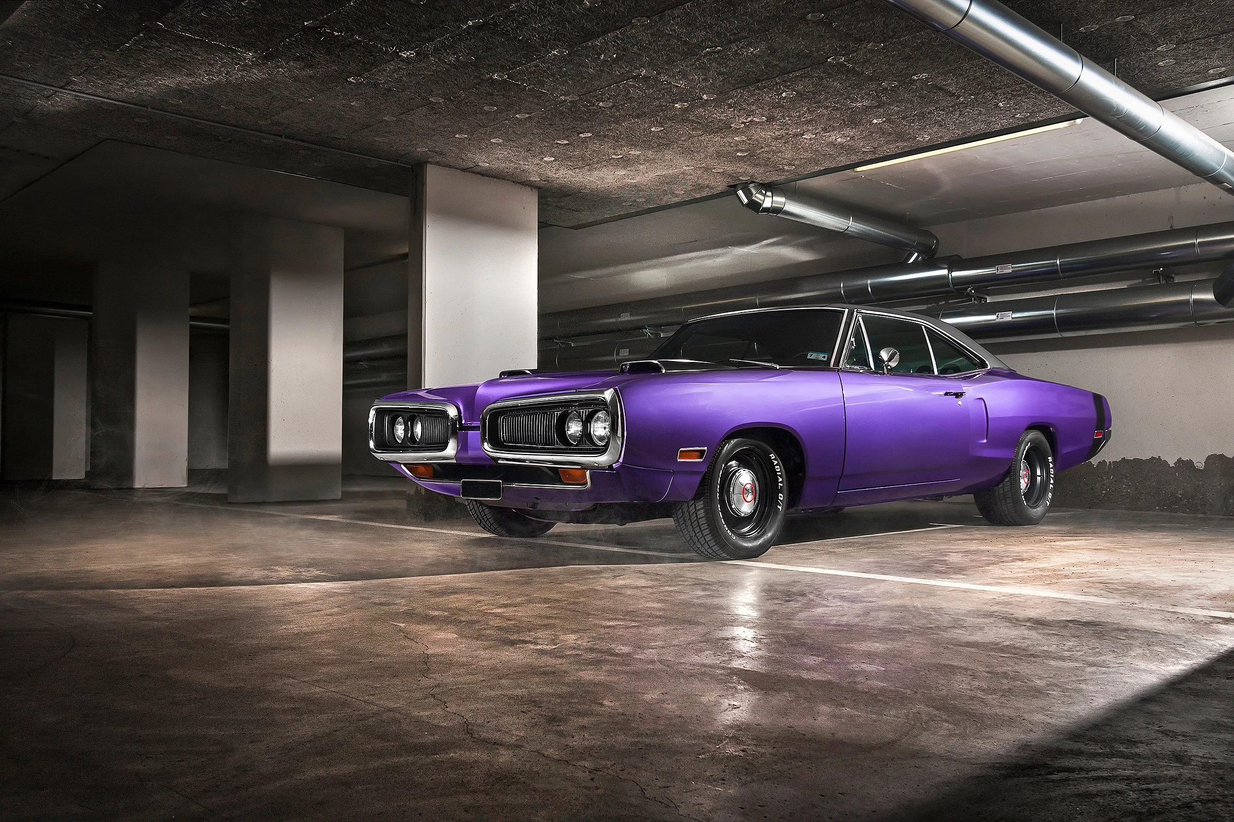 Ein silberner und lila Oldtimer in einer Tiefgarage mit Betonwänden und Decken, angestrahlt von Deckenleuchten.