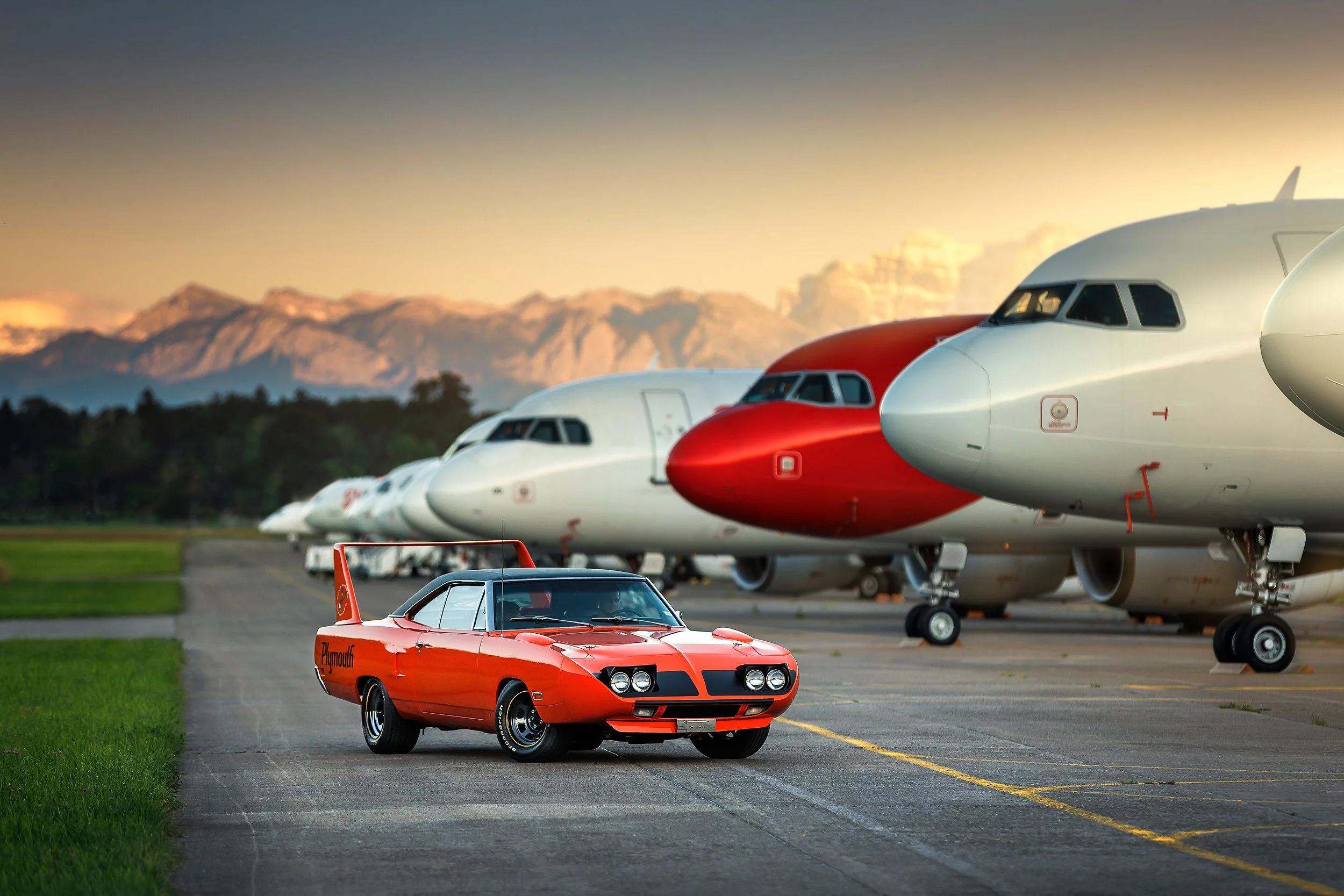 Ein rotes Rennwagen steht auf einem Flughafenvorfeld vor Linienflugzeugen im Hintergrund bei Sonnenuntergang, Berge im Hintergrund.