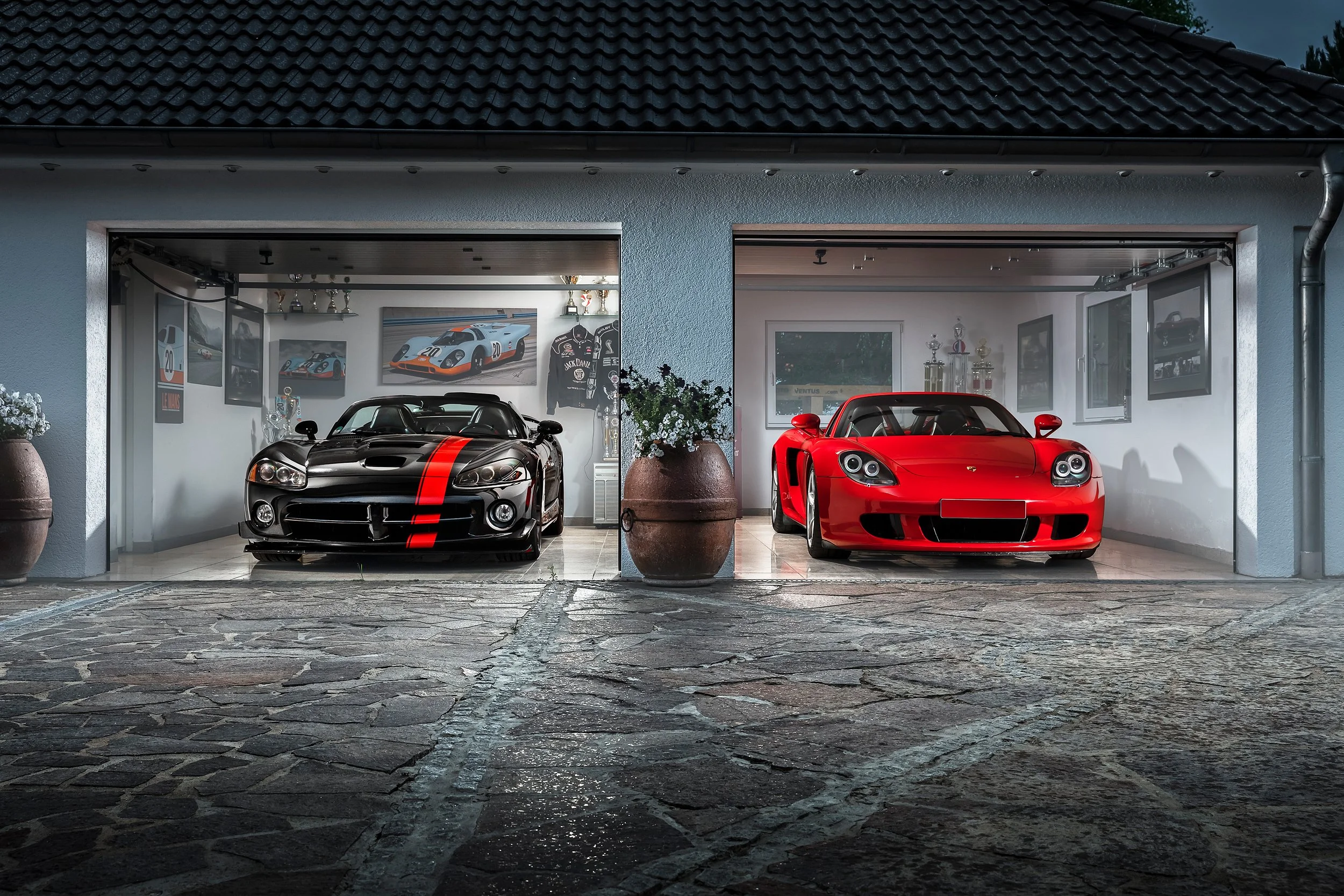 Blick in eine Doppelgaragen mit zwei Sportwagen, einem schwarzen mit rotem Streifen und einem roten, im Inneren an der Wand befinden sich Rennsport-Poster, Trophäen und ein Jack Daniel's Hemd.