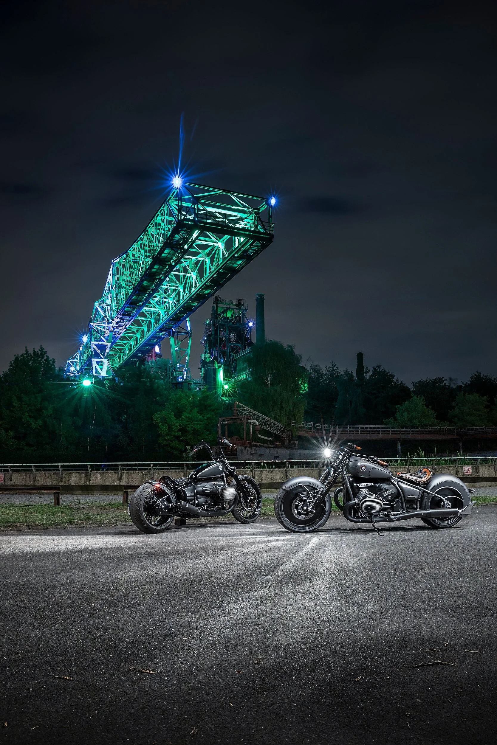 Zwei schwarze Motorräder stehen nebeneinander auf einer Straße vor einer Lagerhalle, im Hintergrund leuchtende Industrieanlage mit blauen und grünen Lichtern, nachts, dunkler Himmel, im Vordergrund Straße und einige Blätter auf dem Boden.