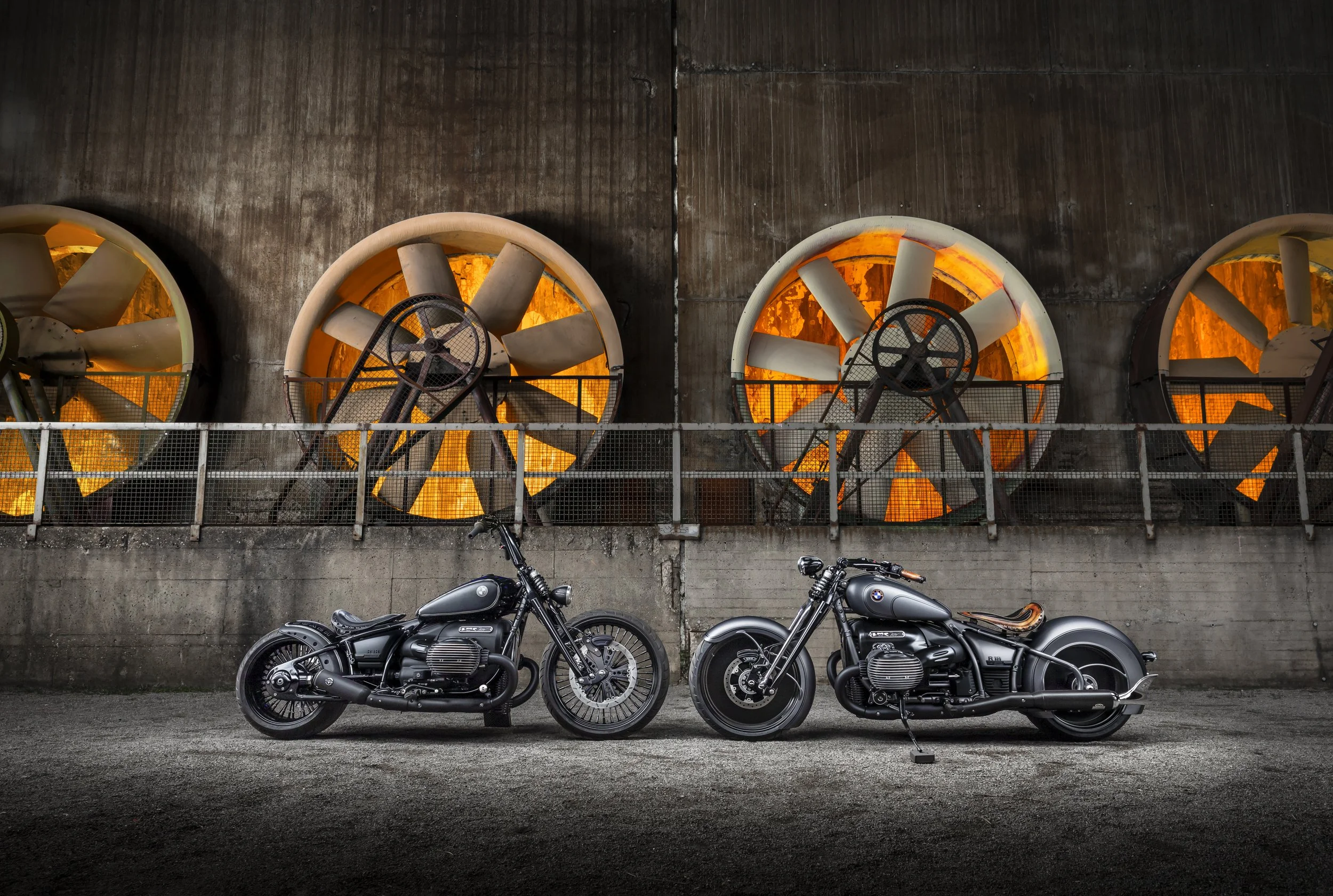 Zwei schwarze Motorräder vor einer Betonwand mit großen Ventilatoren, die orange leuchten, und Fahrrädern im Vordergrund.