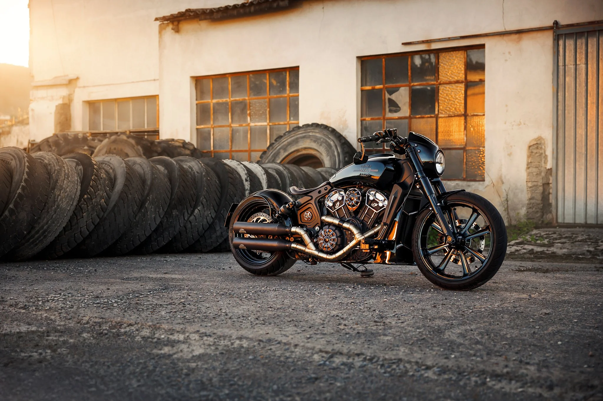 Eine schwarze Harley-Davidson Scout Motorrad steht vor einem alten, mit Reifen und Autoteilen gefüllten Lagerhaus bei Sonnenuntergang.