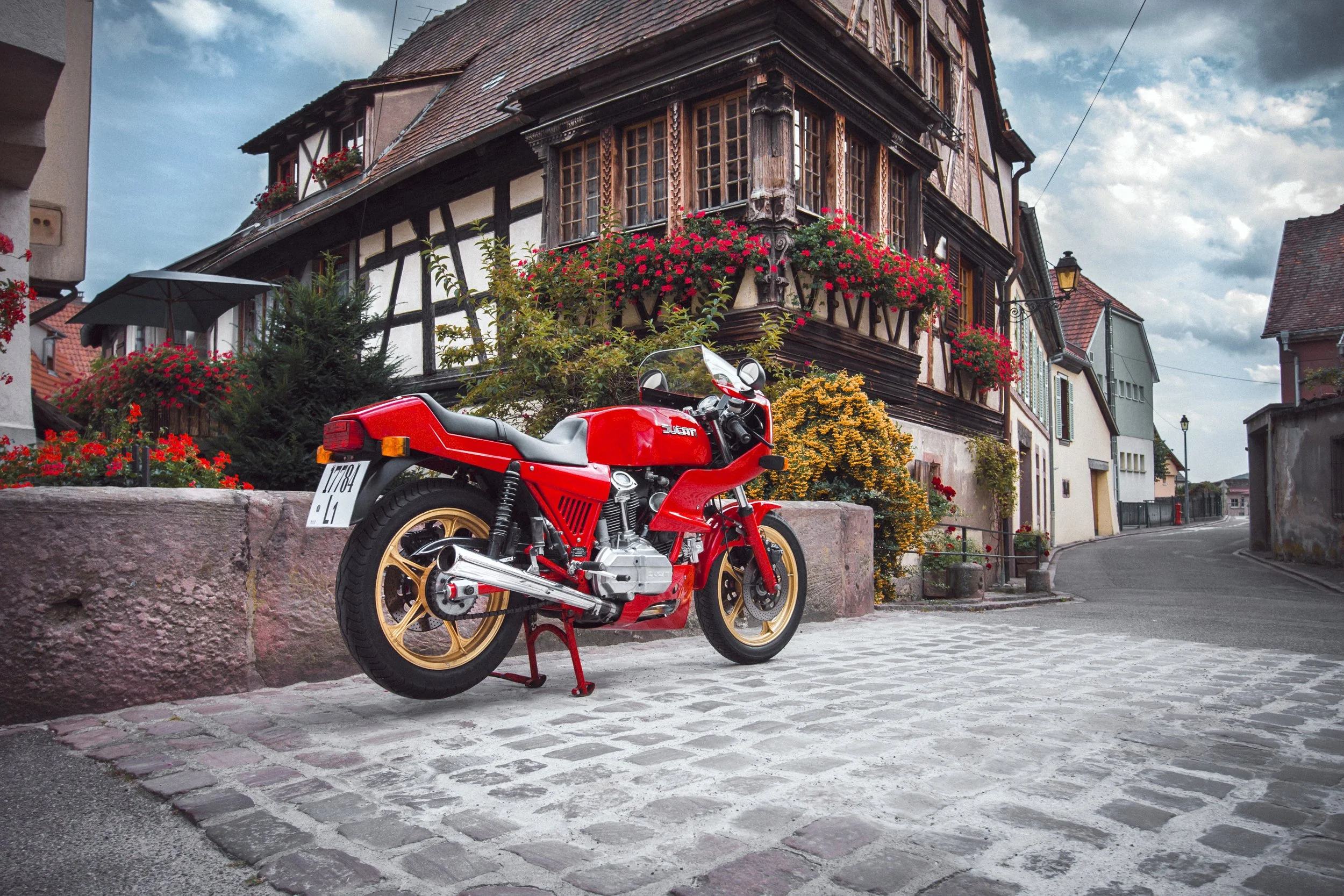 Rote Ducati Motorrad vor einem historischen Fachwerkhaus mit Blumen im Fenster und Straßenlaterne in einer kleinen europäischen Stadt an einem bewölkten Tag.