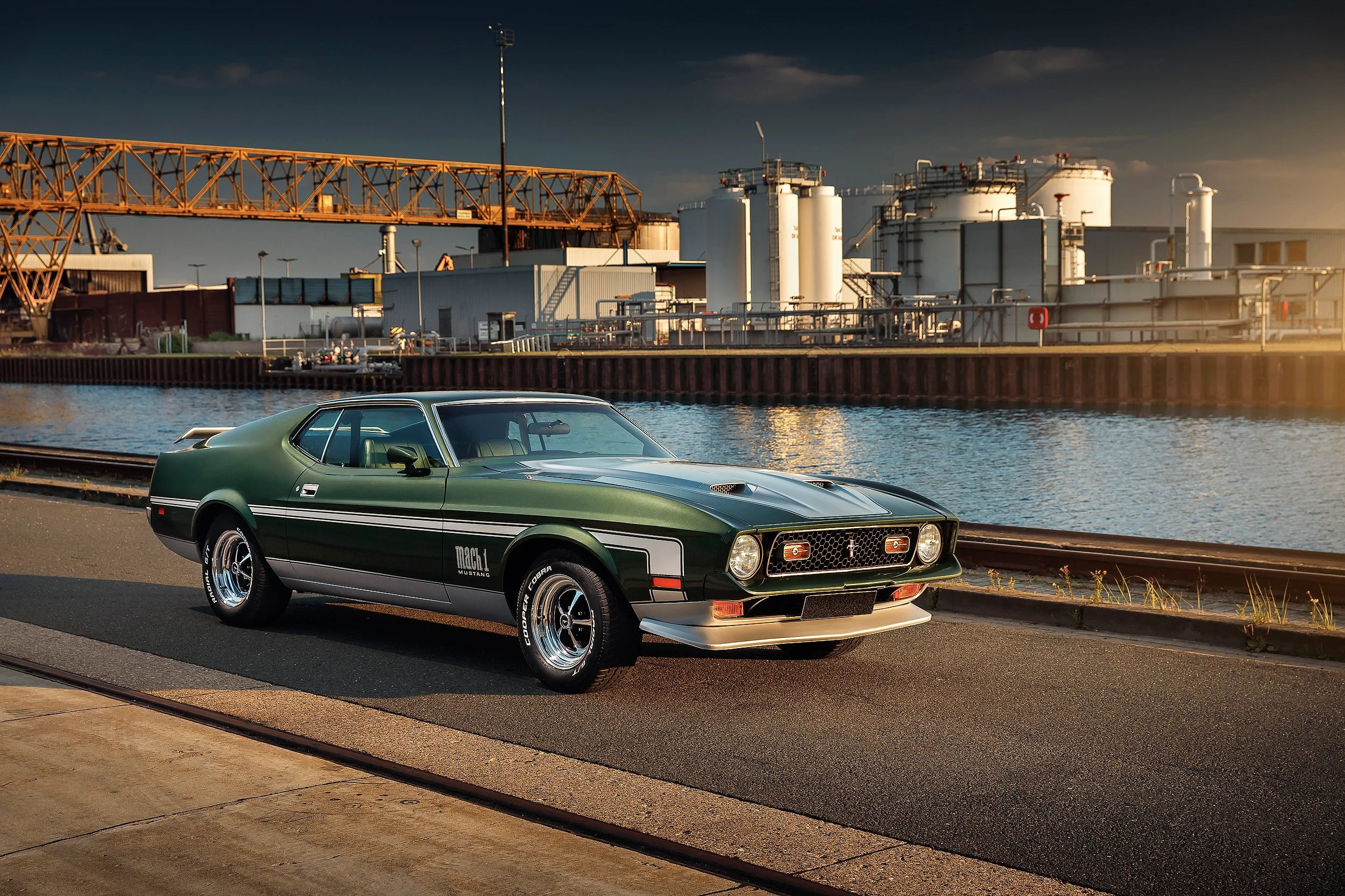 Ein grüner Mustang Mach 1 Oldtimer-Pferdeauto steht am Wasser entlang, im Hintergrund eine Industrieanlage mit Tanks und einer Brücke bei Sonnenuntergang.