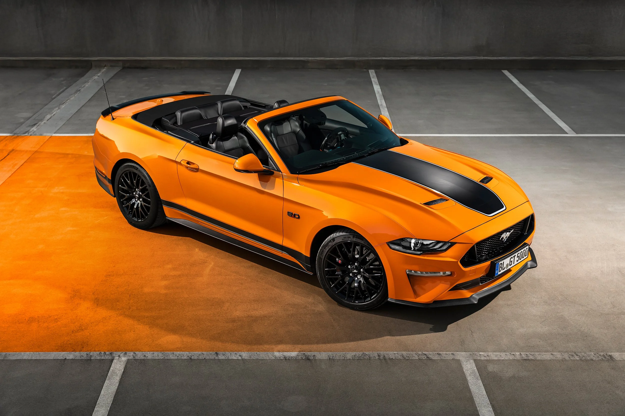Ein orangefarbener Cabrio, ein Ford Mustang, steht in einer leeren Parkgarage auf einer orangefarbenen Matte.
