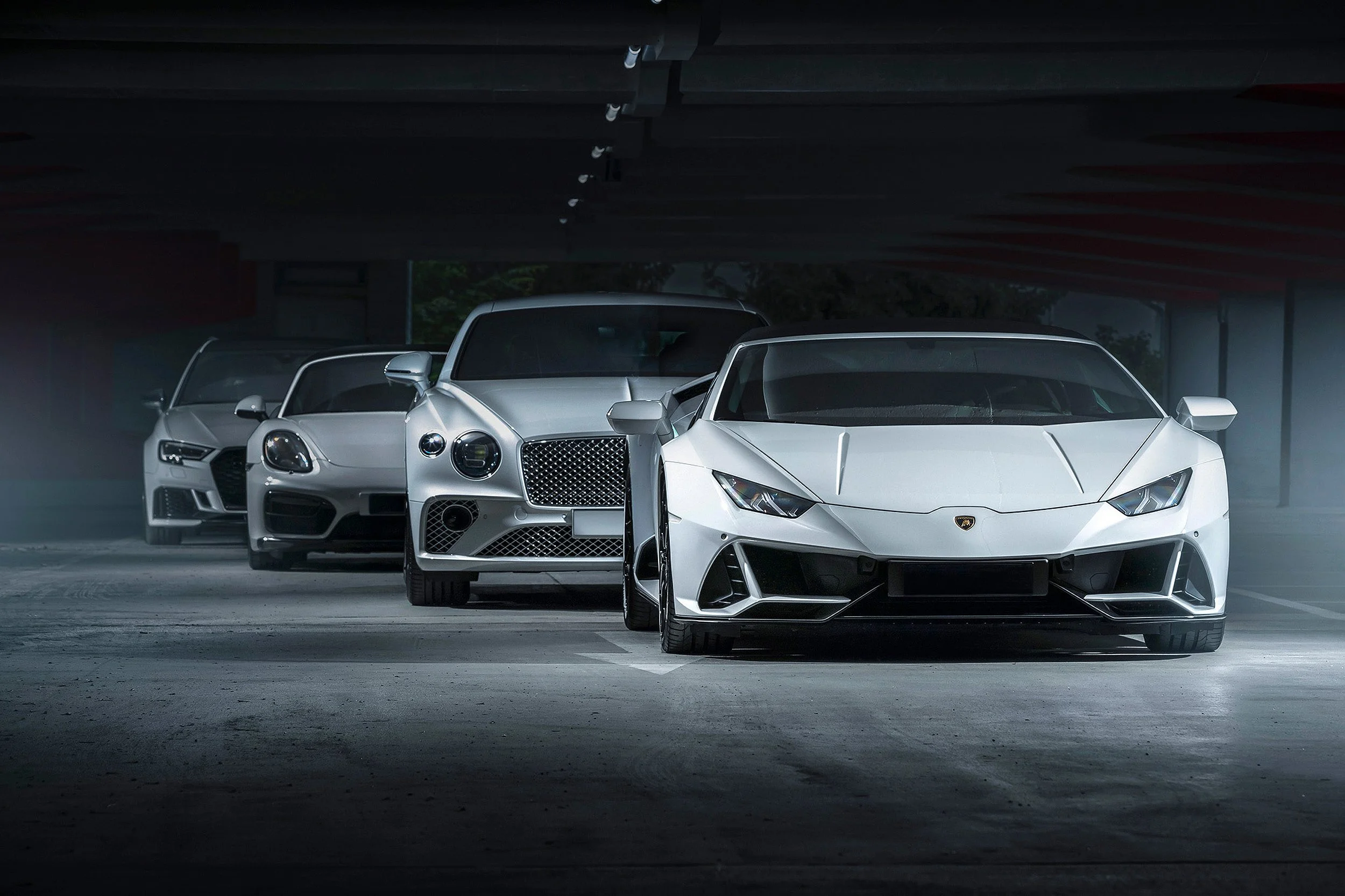 Verschiedene Luxusautos, darunter ein weißer Lamborghini, stehen in einer dunklen Tiefgarage.