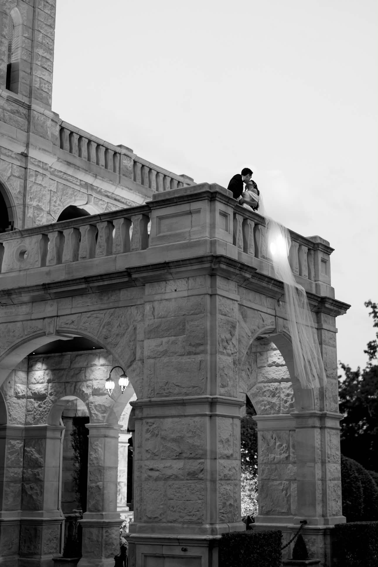 A black-and-white photo of a stone building with arches and decorative columns. A couple gently leaning in for a kiss is on the upper balcony, with a flowing curtain hanging from the railing.