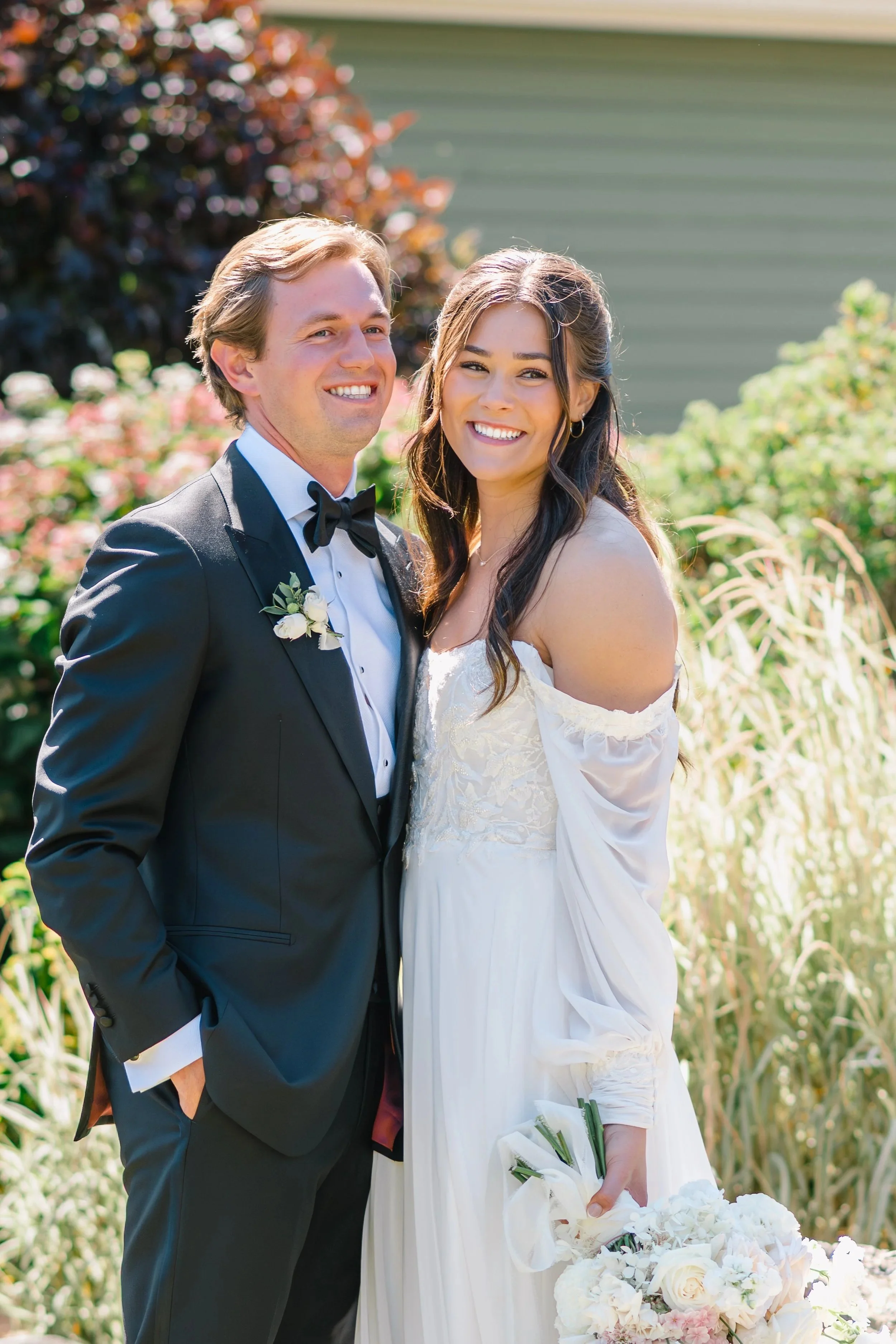 A newlywed couple standing outdoors, smiling, with the groom in a black tuxedo and the bride in a white wedding dress holding a bouquet of white and pink flowers.