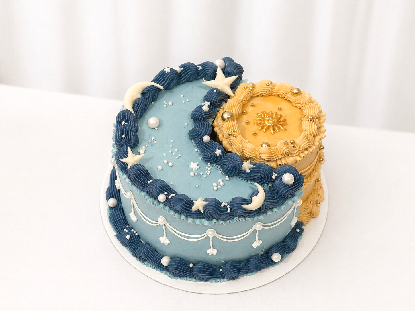 A celestial-themed cake with blue and gold frosting, decorated with stars, moons, a sun emblem, and pearl-like ornaments.