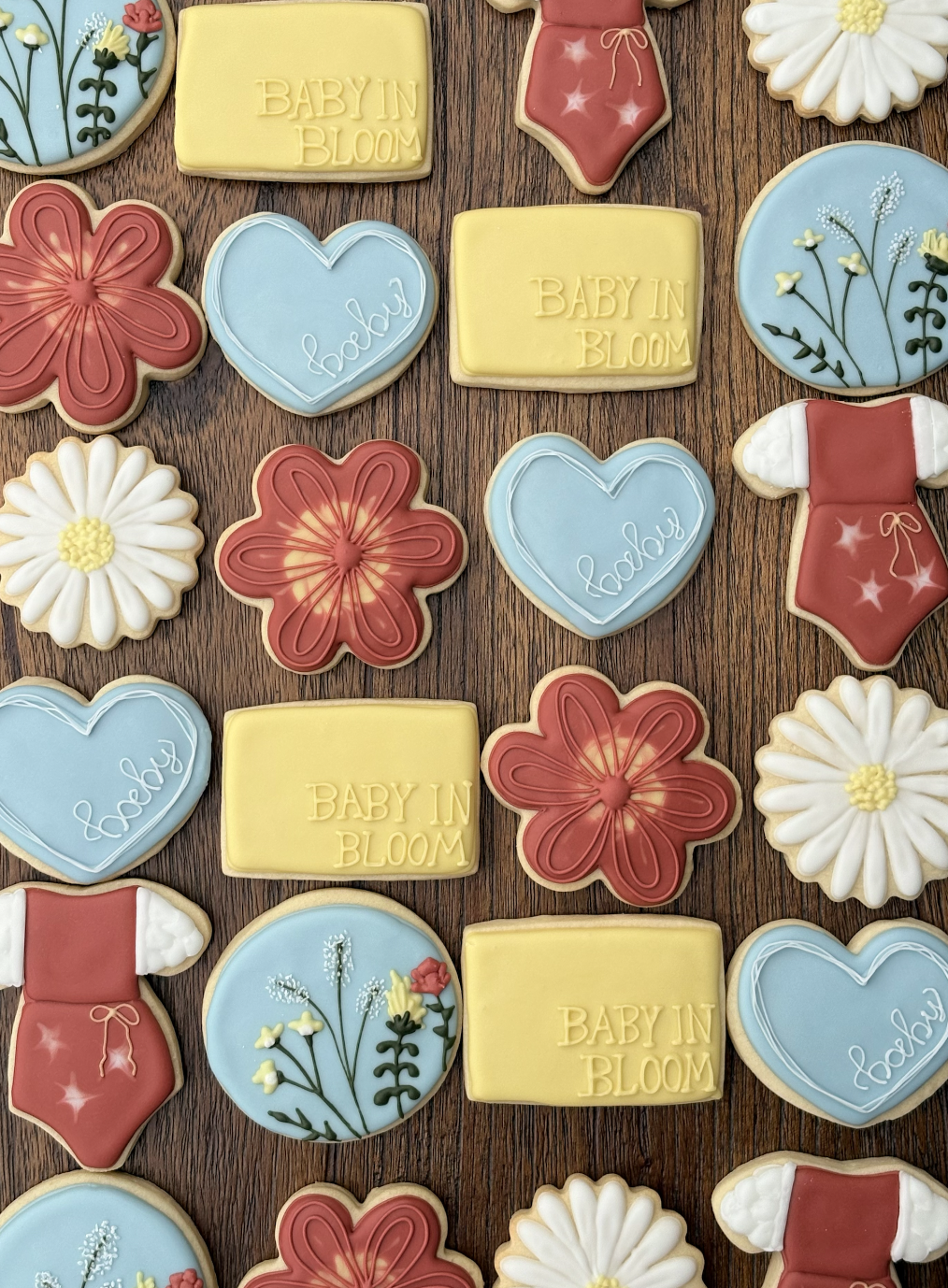 Decorative cookies shaped like flowers, hearts, and baby clothes, with pastel colors and inscriptions like 'Baby in Bloom' and 'honey', arranged on a wooden surface.