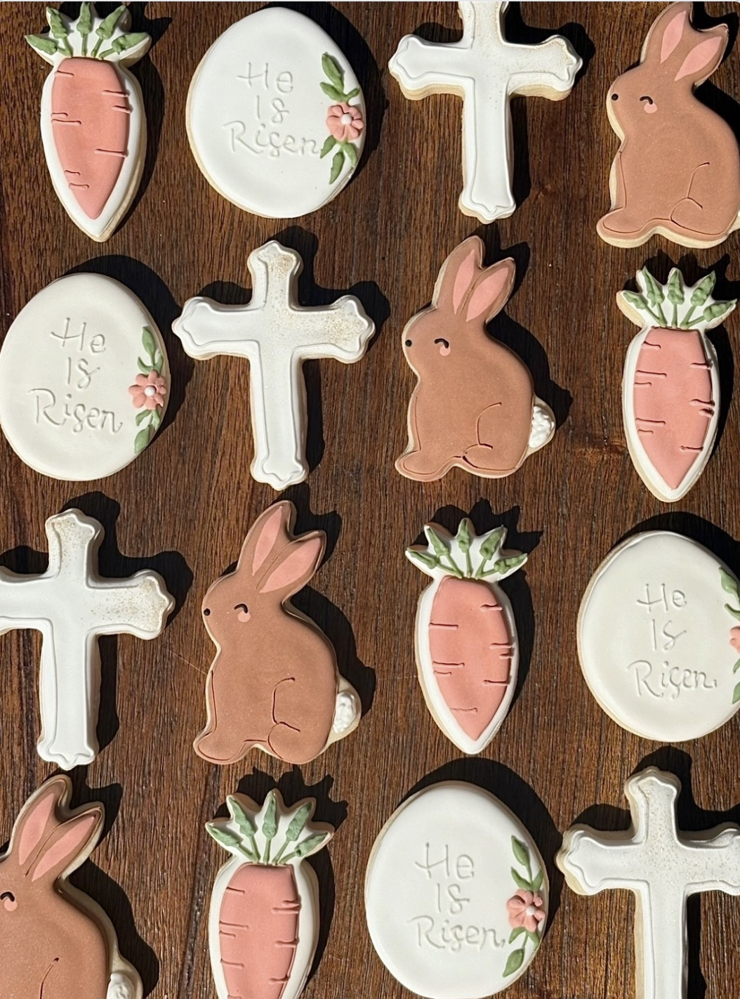 Decorative cookies shaped like carrots, rabbits, crosses, and eggs with the message 'He Is Risen' on some eggs, arranged on a wooden surface for Easter.