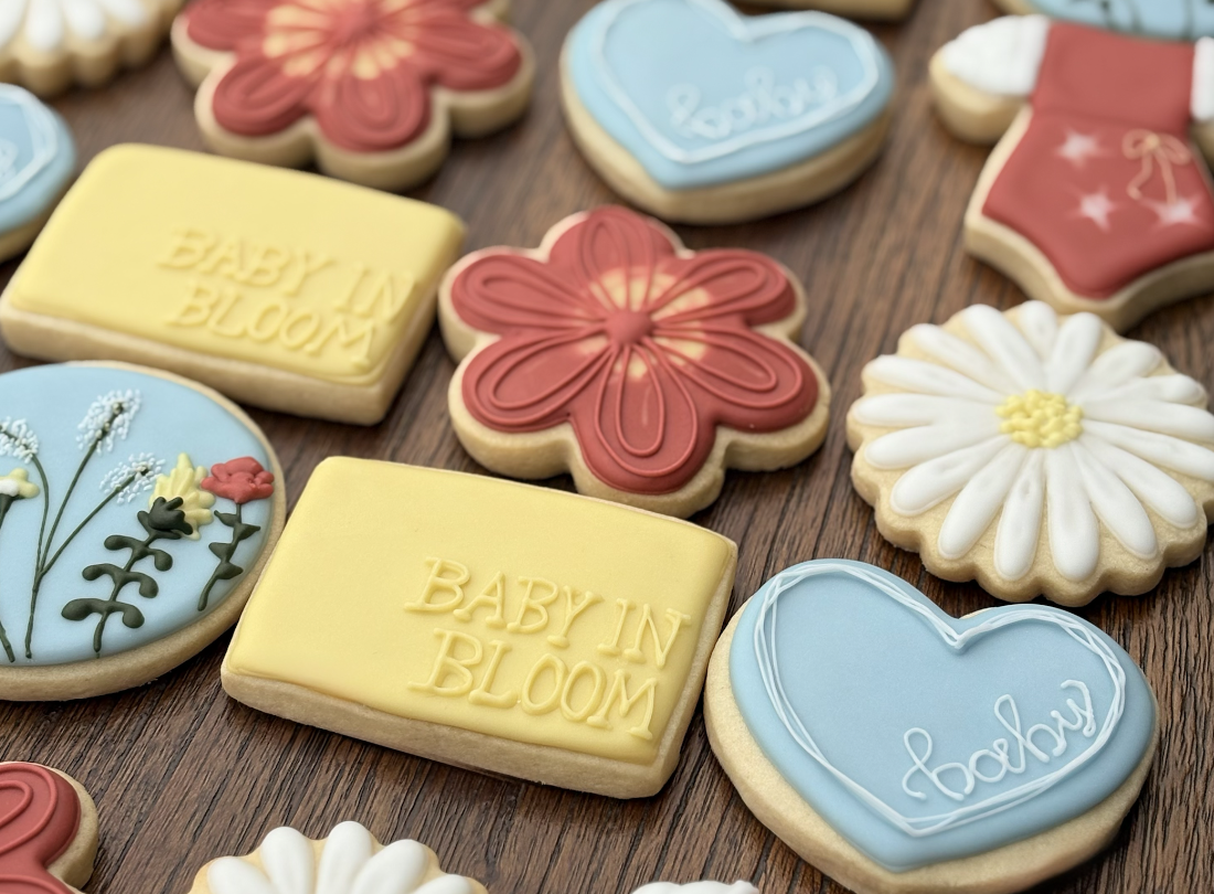 Assorted decorated cookies on a wooden surface, including flowers, hearts, and books with messages like "Baby in Bloom" and "Peekaboo".