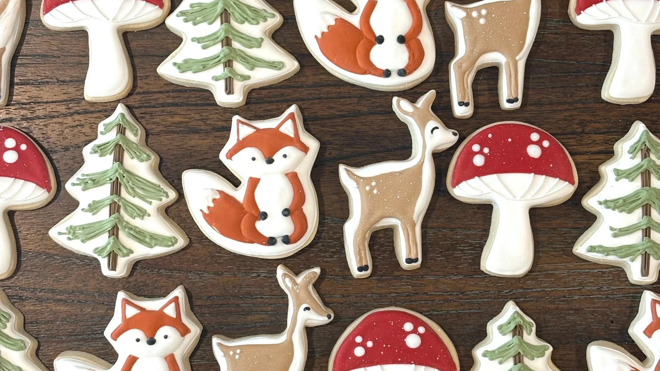 Decorated holiday cookies shaped like mushrooms, pine trees, foxes, fox faces, and deer on a wooden surface.