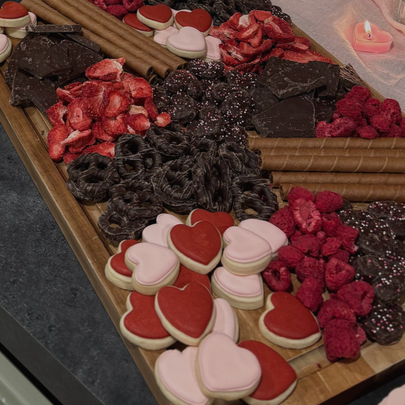 Assorted Valentine's Day cookies, chocolates, and dried strawberries on a wooden tray next to a lit pink candle.