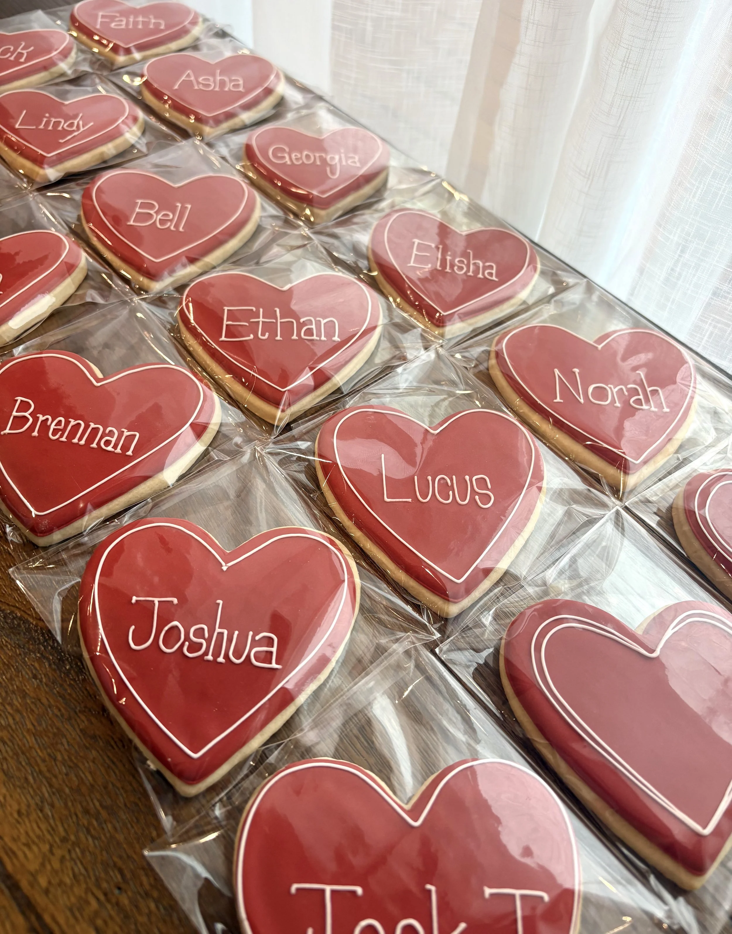 Heart-shaped decorated cookies with red icing, each with a child's name written in white icing, arranged on a table and wrapped in clear plastic.