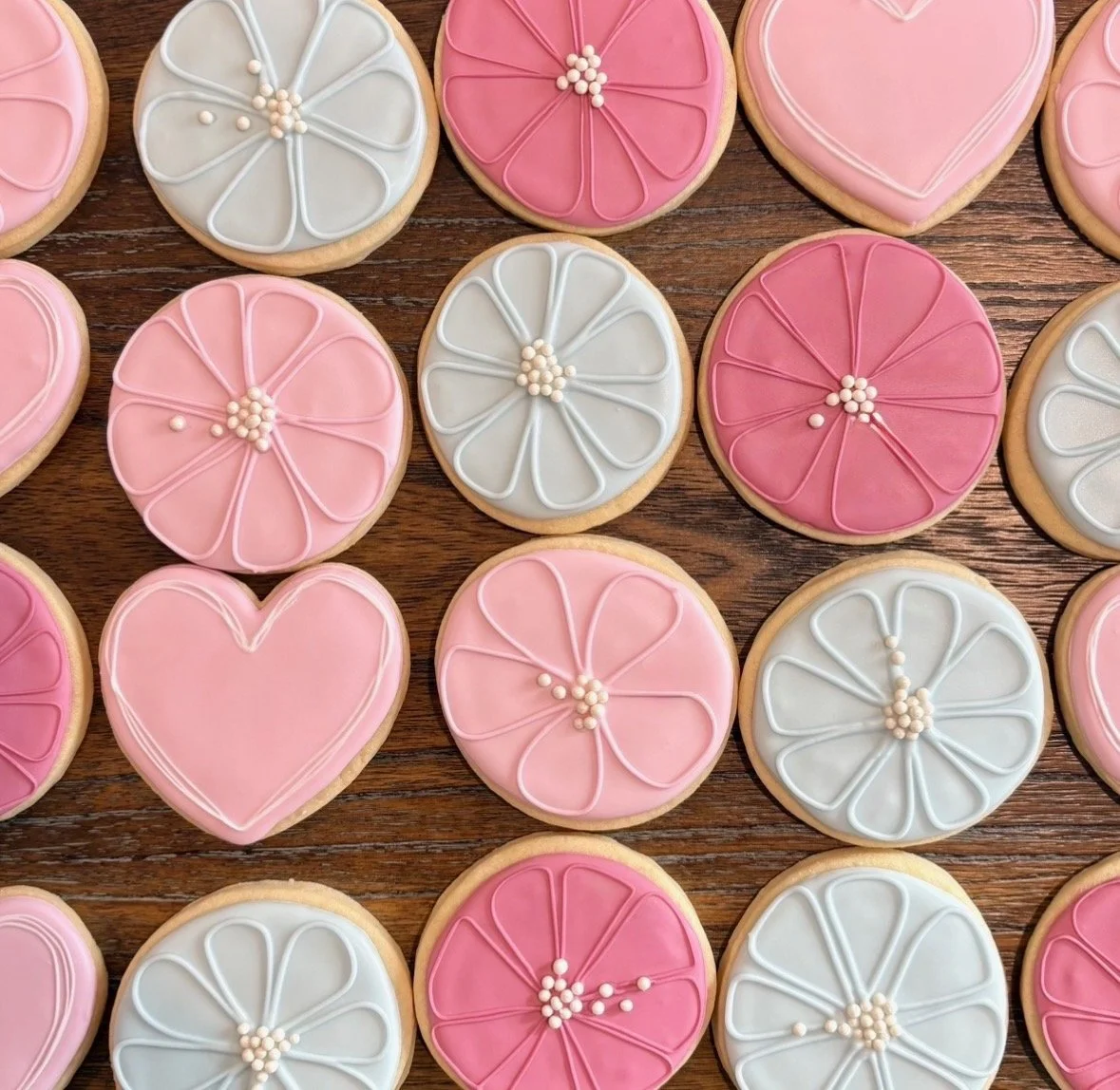 Decorated sugar cookies in pink, white, and light blue with floral and heart designs, arranged on a wooden surface.