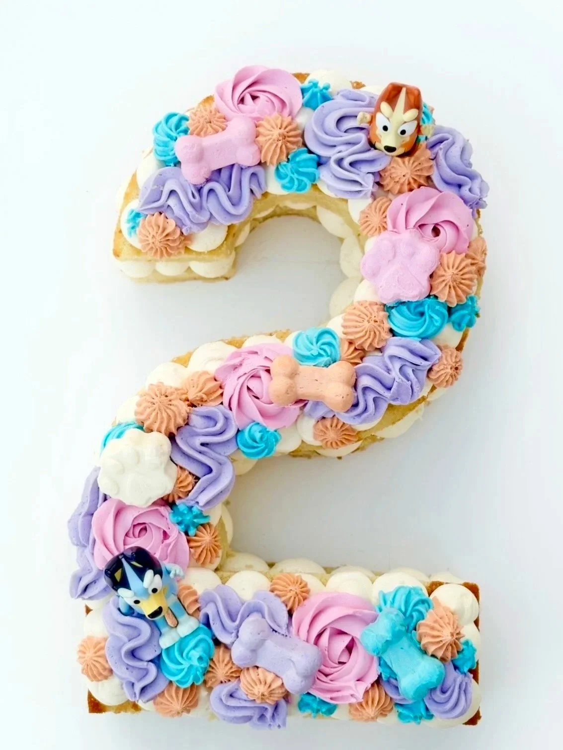 Decorated birthday cake in the shape of the number 2, with pastel-colored frosting, small toy characters, and bone-shaped decorations.