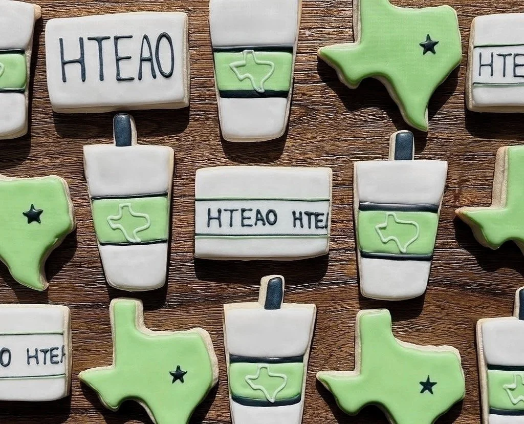 Cookies shaped like Texas, coffee cups, and blocks with the word 'HTEAO' written on them, decorated in green, white, and black icing, arranged on a wooden surface.