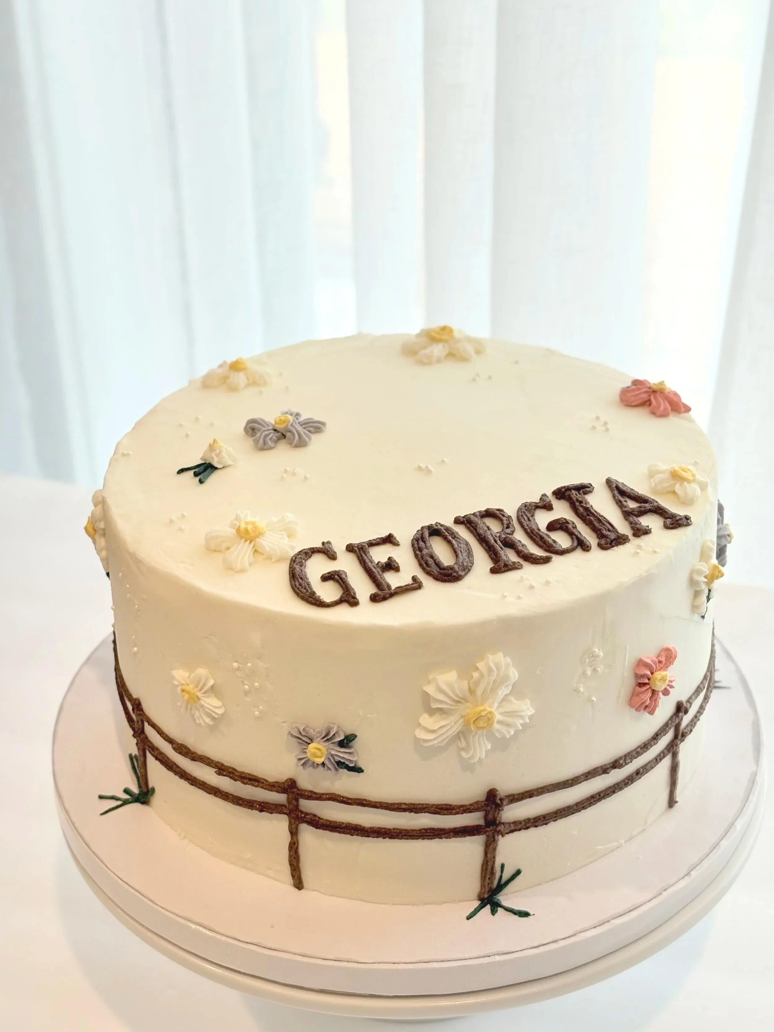 A round white cake decorated with small pastel-colored flower-shaped icing decorations and the word 'GIDEON' written in chocolate on the top. The cake has a chocolate rope border around the middle and bottom edges, and is placed on a white cake stand.
