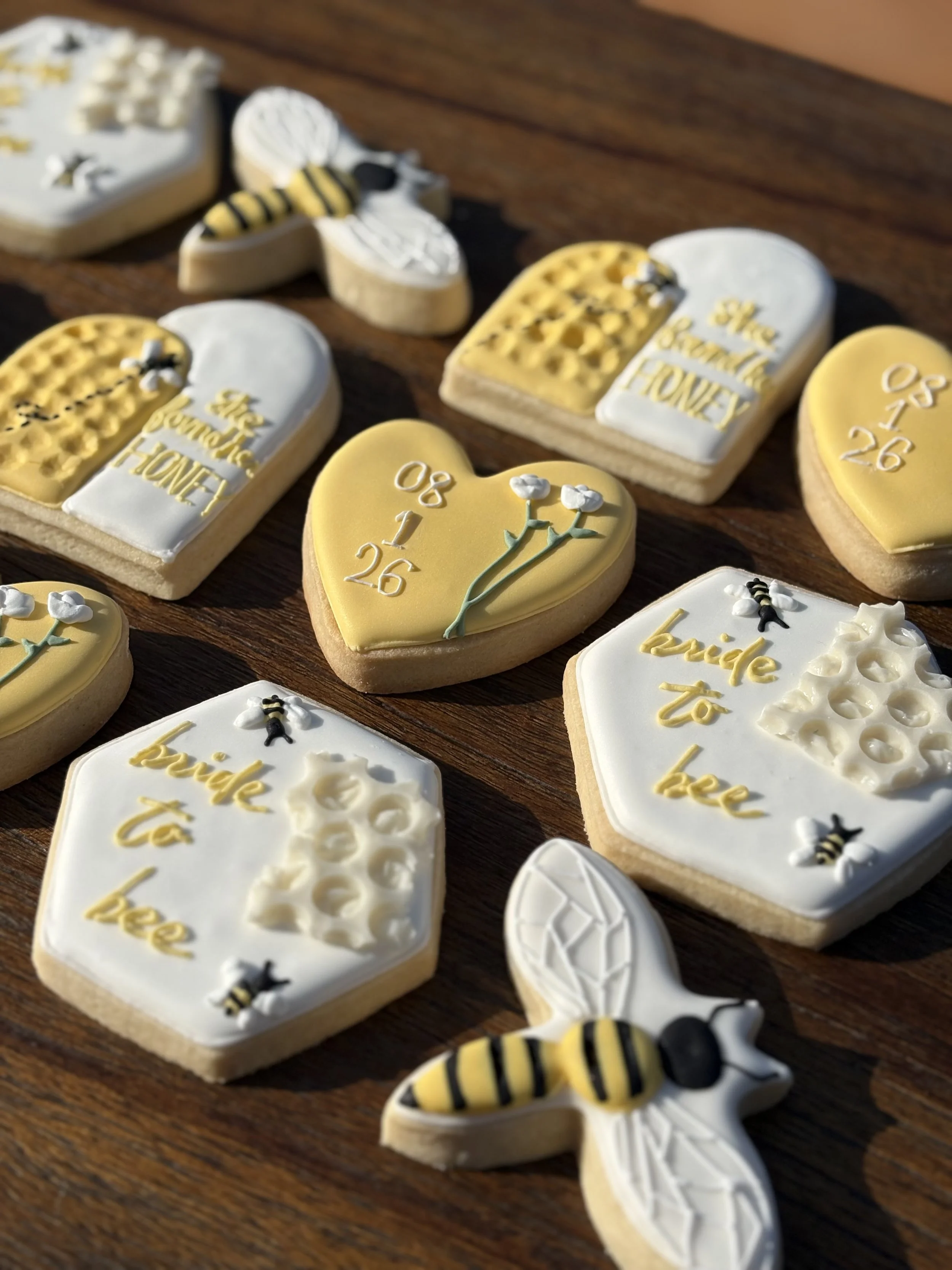 Decorated cookies with bee and honey themes, some reading 'Bee kind' and 'She believed she could so she did', on a wooden surface.