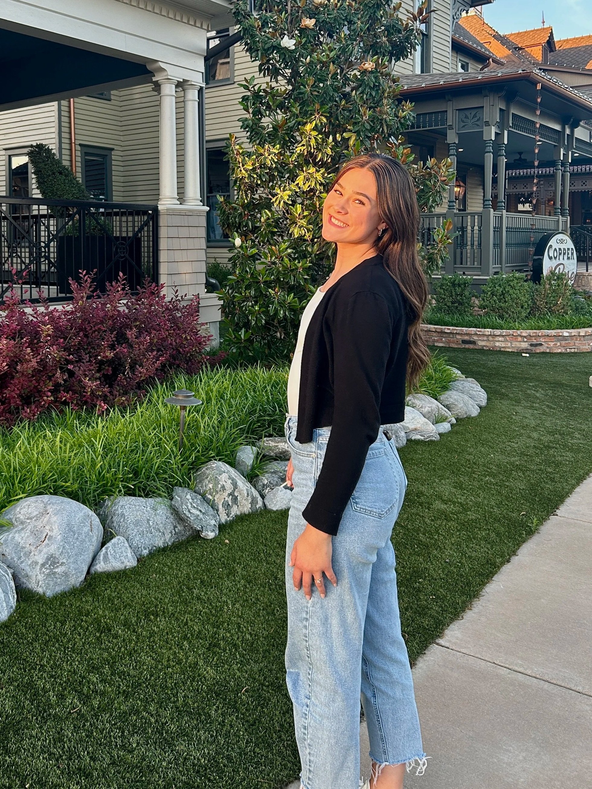 A woman standing outdoors on a sidewalk in front of a house with garden, smiling with hair flowing, wearing a black jacket and jeans.
