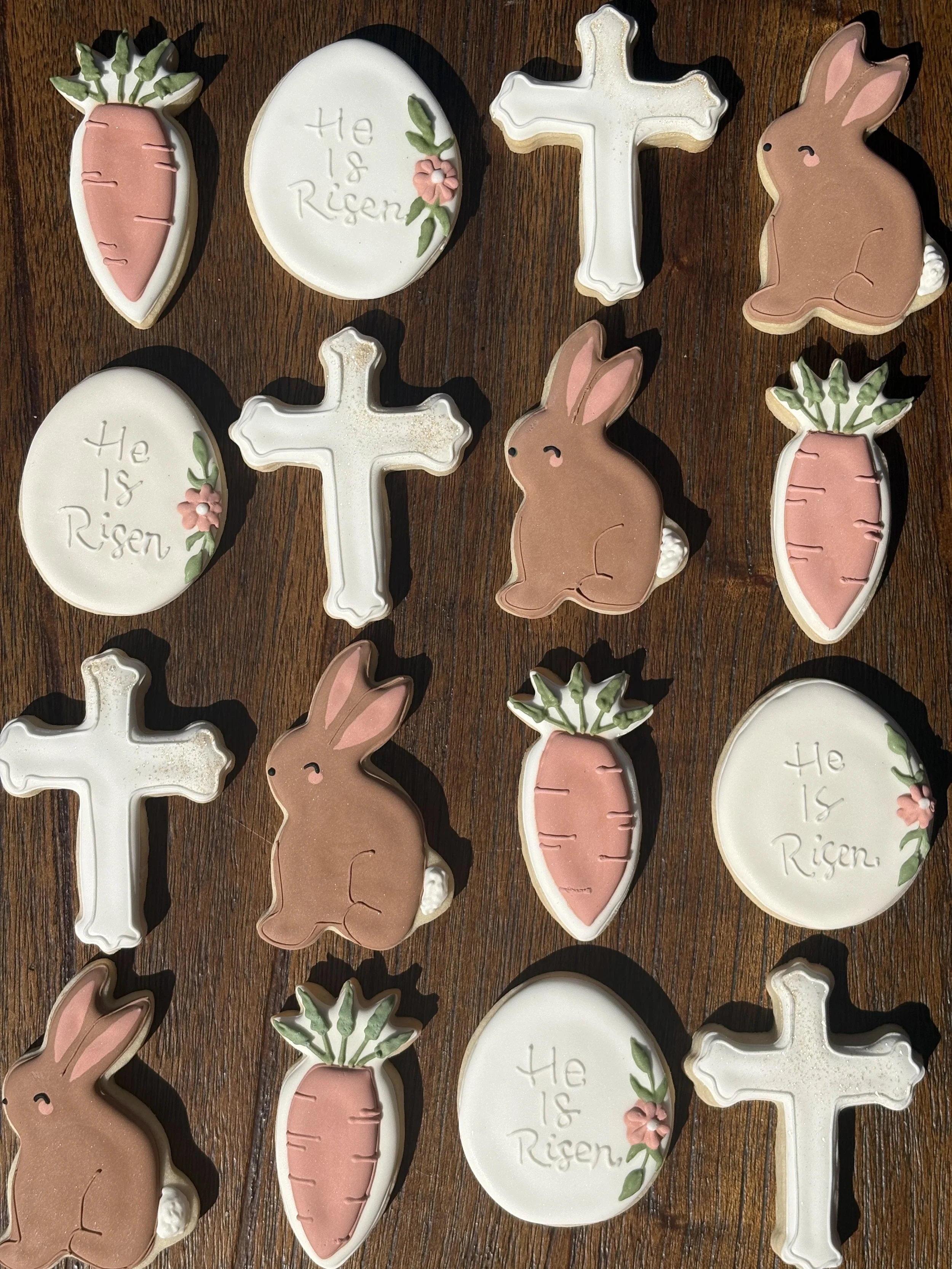 Assorted decorated cookies on a wooden surface including carrots, crosses, rabbits, and round cookies with 'He is Risen' written and floral designs.