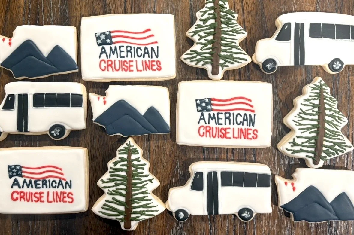 Decorated cookies with American cruise line and travel themes, including trees, mountains, and buses, on a wooden surface.