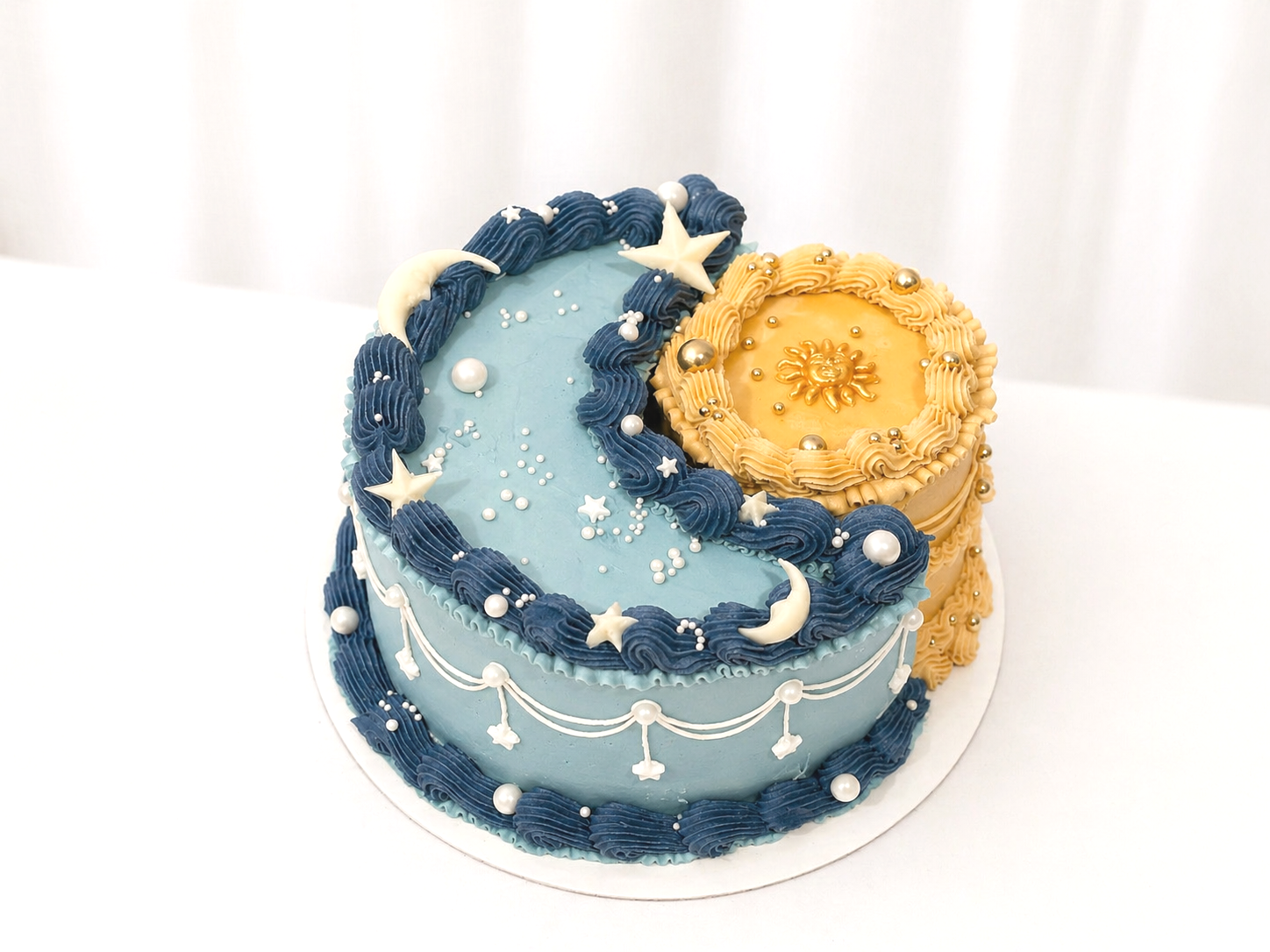 Decorative birthday cake with celestial theme, featuring blue and gold icing, stars, moons, pearls, and a sun emblem on top.