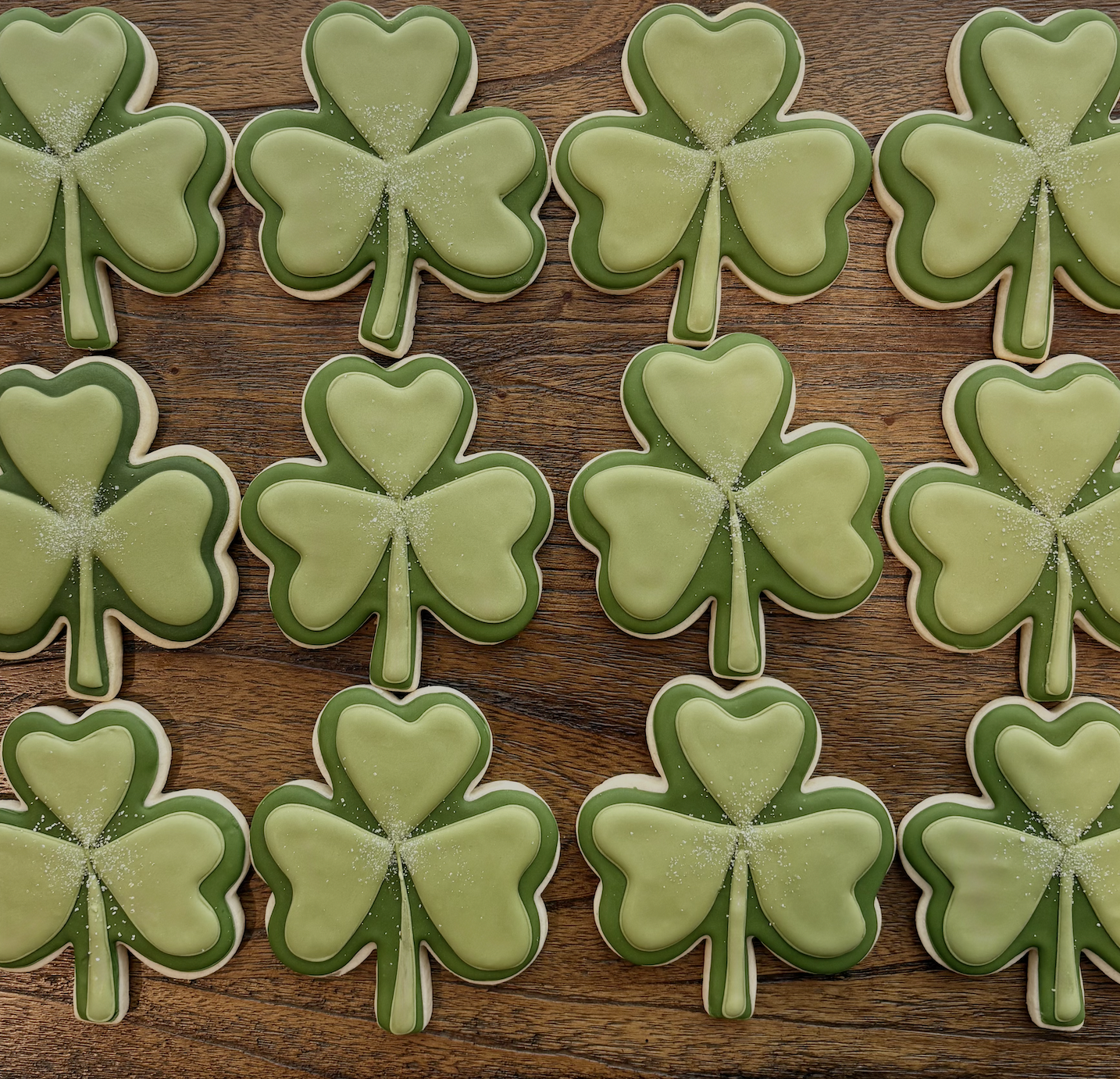 Green shamrock-shaped cookies decorated with green icing and white sugar on a wooden surface.