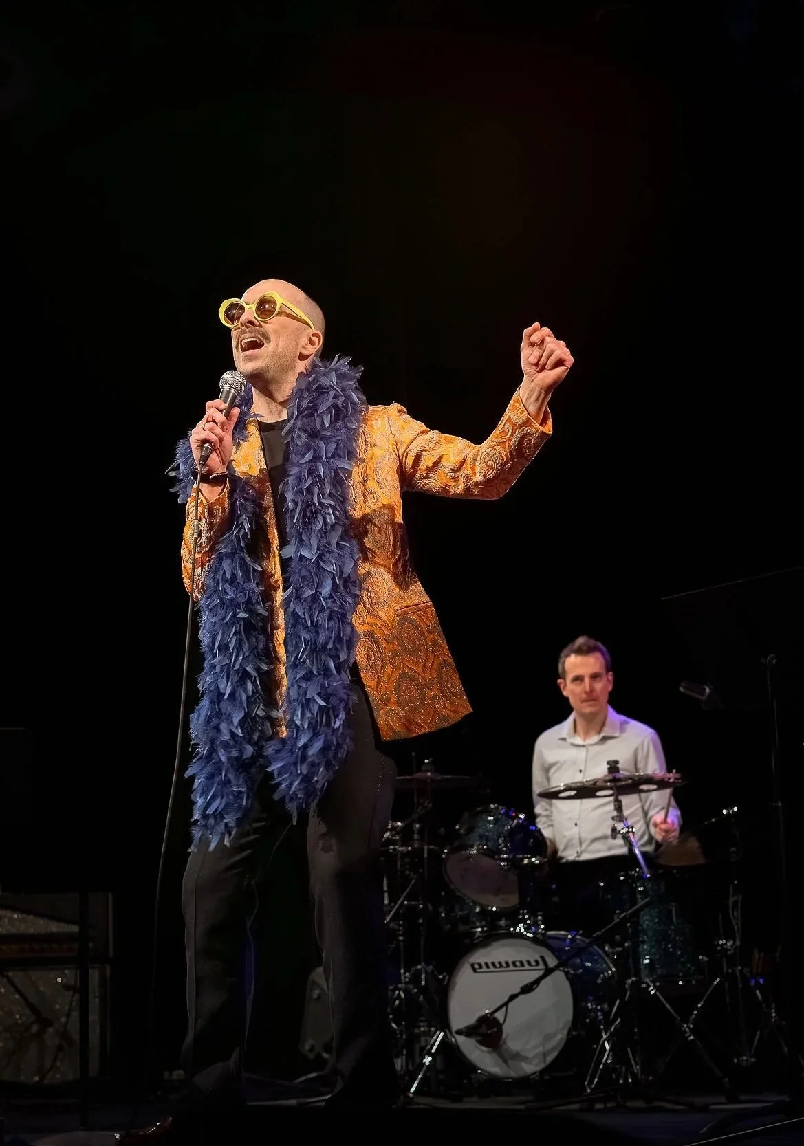 Aaron Hutton with yellow sunglasses singing into a microphone on stage, wearing an orange patterned jacket and a blue feathered scarf, with a drummer in the background.