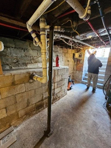 Person standing in a dim unfinished basement with exposed plumbing, electrical wiring, cinder block walls, and a fire extinguisher