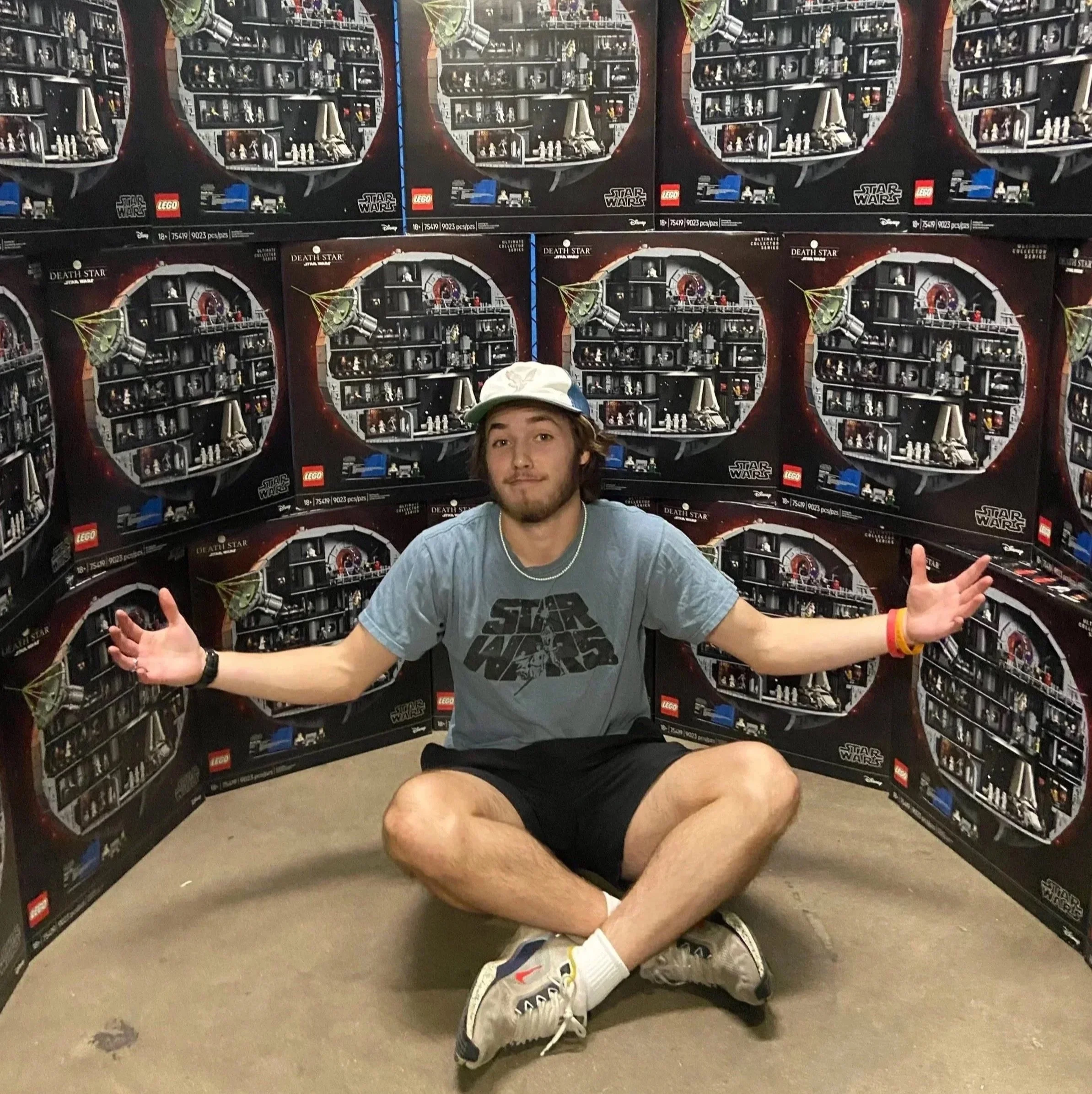 A young man sitting on the floor with his back against a wall of Star Wars LEGO sets, with his arms wide open and a relaxed expression.