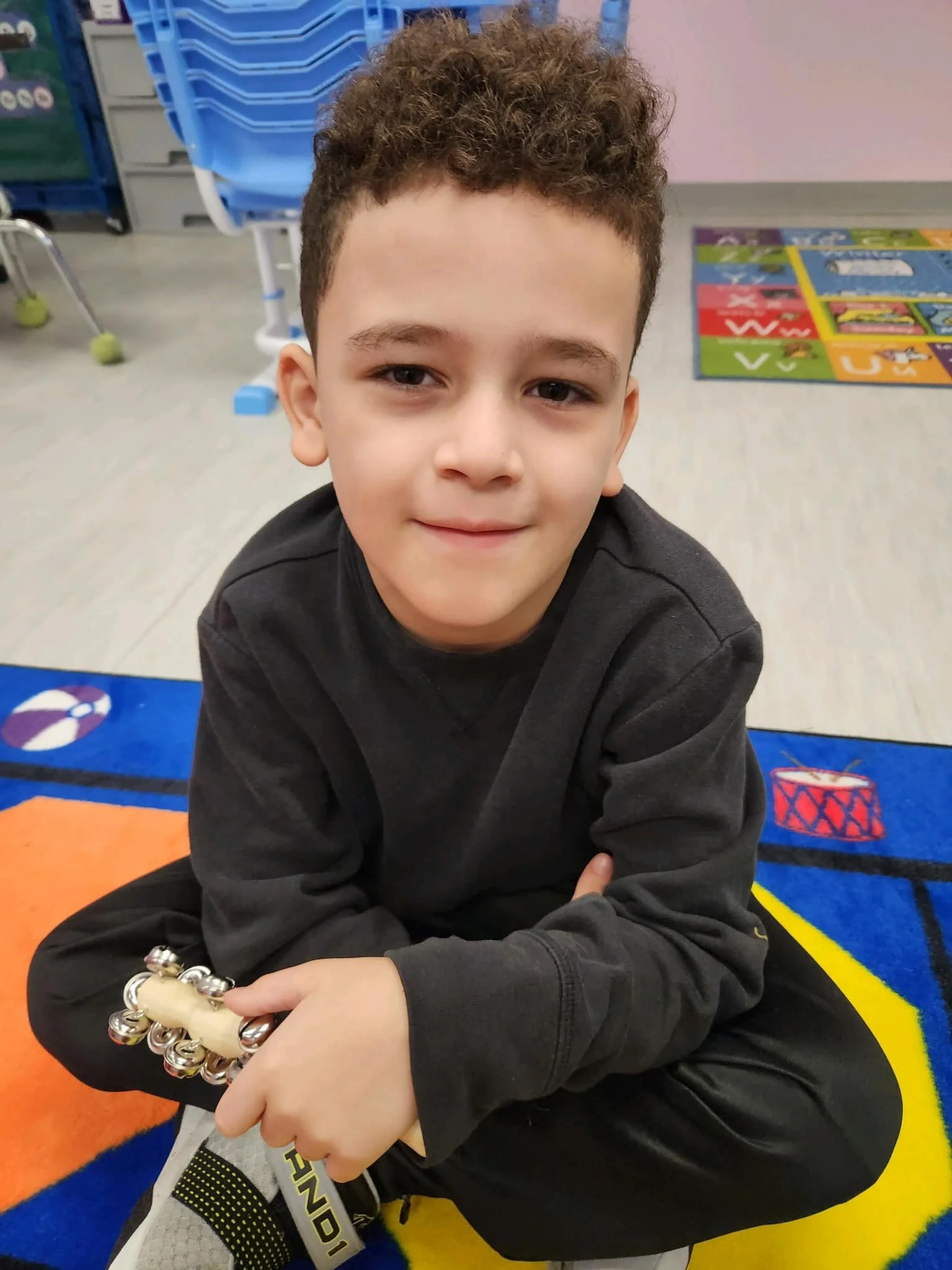 A young boy with short curly hair, wearing a black long-sleeve shirt, sitting on a colorful children's rug in a classroom, holding a small musical instrument, with classroom furniture and educational materials in the background.